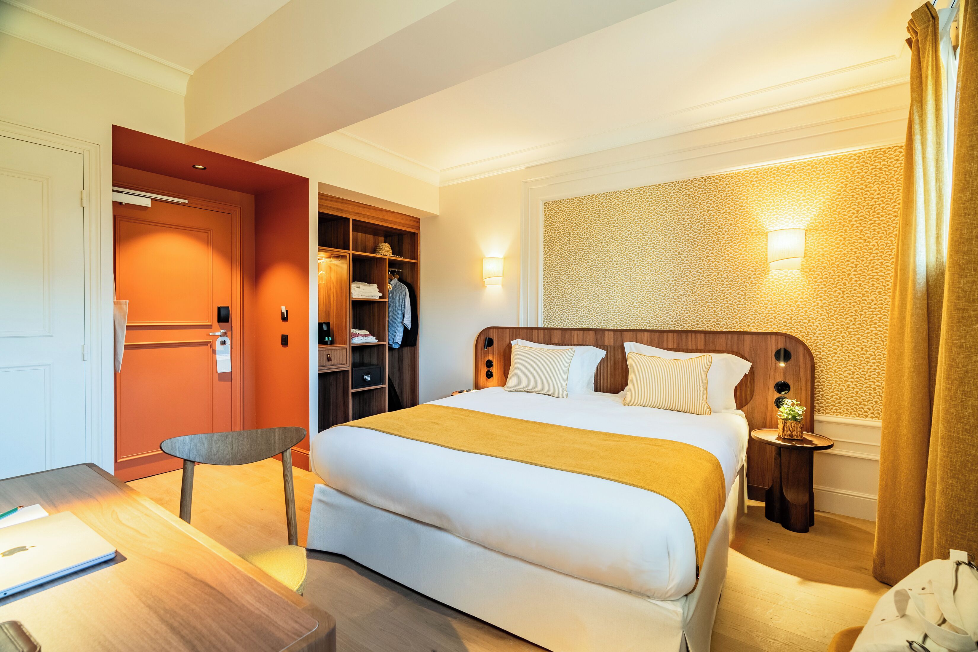 A hotel room with a large bed, a desk with a laptop, a wardrobe, and a small table with a plant.