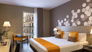 A hotel room with a large bed, a desk with a chair, a window with a view of buildings, and a wall with a geometric pattern. - Citadines Ramblas Barcelona (Barcelona)