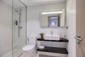 Un baño moderno con ducha envuelta en vidrio, inodoro blanco, encimera negra con lavamanos y espejo sobre el lavamanos.