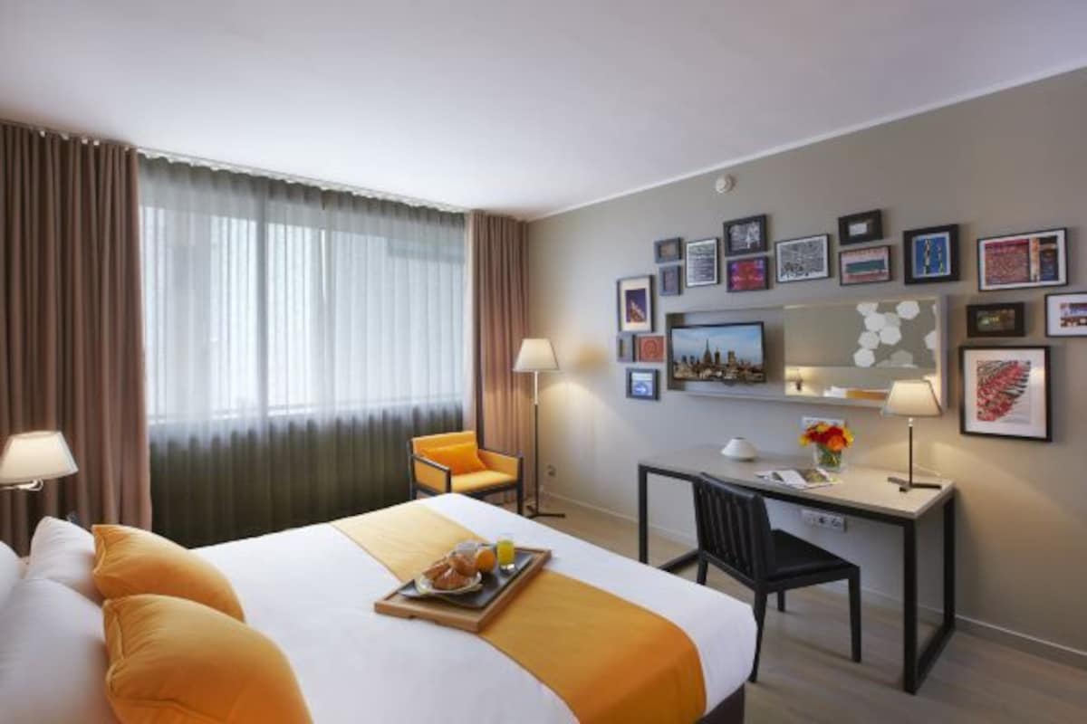A hotel room with a bed, a desk, a chair, a dining table with breakfast, and a wall with framed pictures.