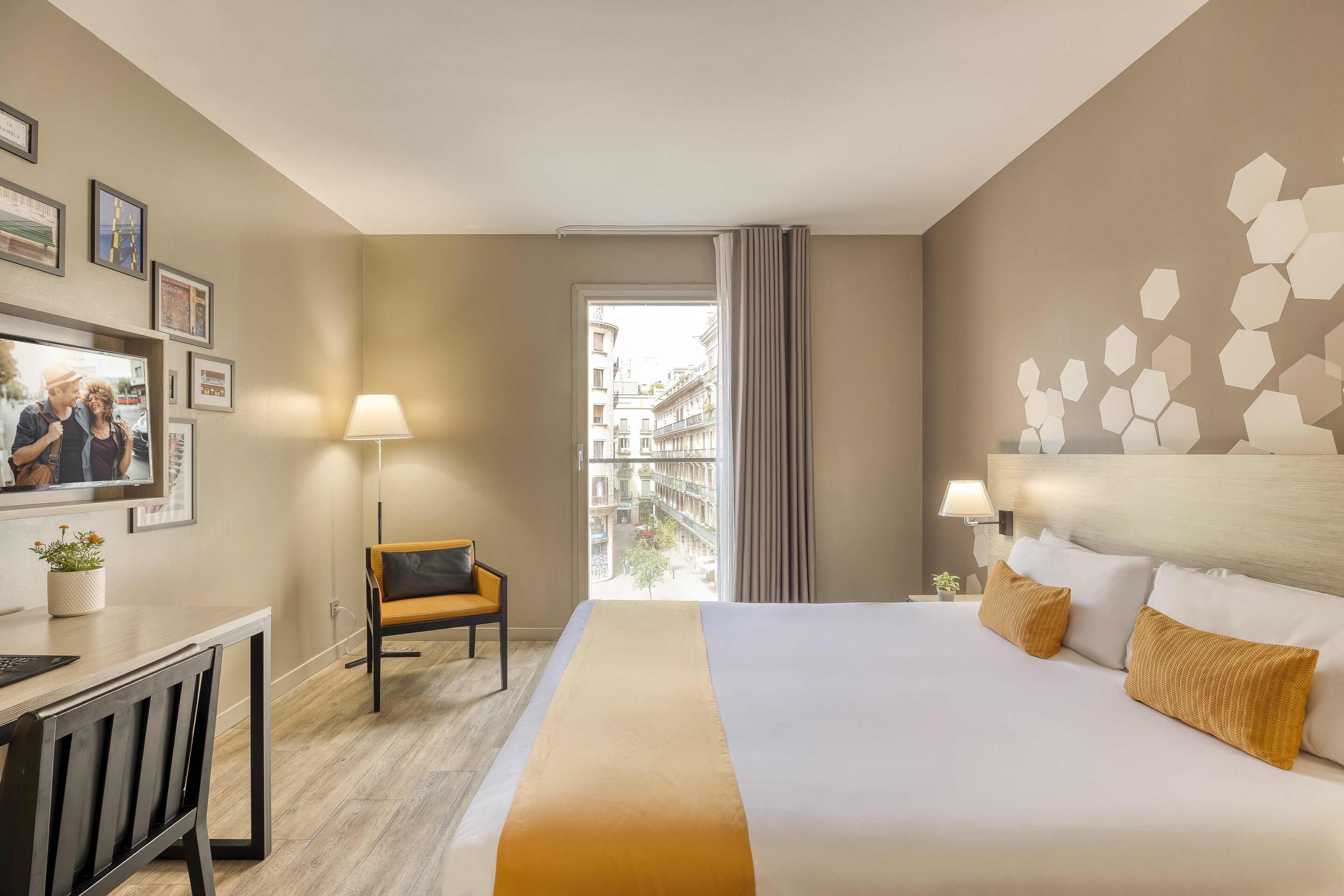 A hotel room with a bed, a desk, a chair, a lamp, and a view of the city.