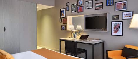 Quarto de hotel com cama, escrivaninha, cadeira, televisão e obras de arte nas paredes.