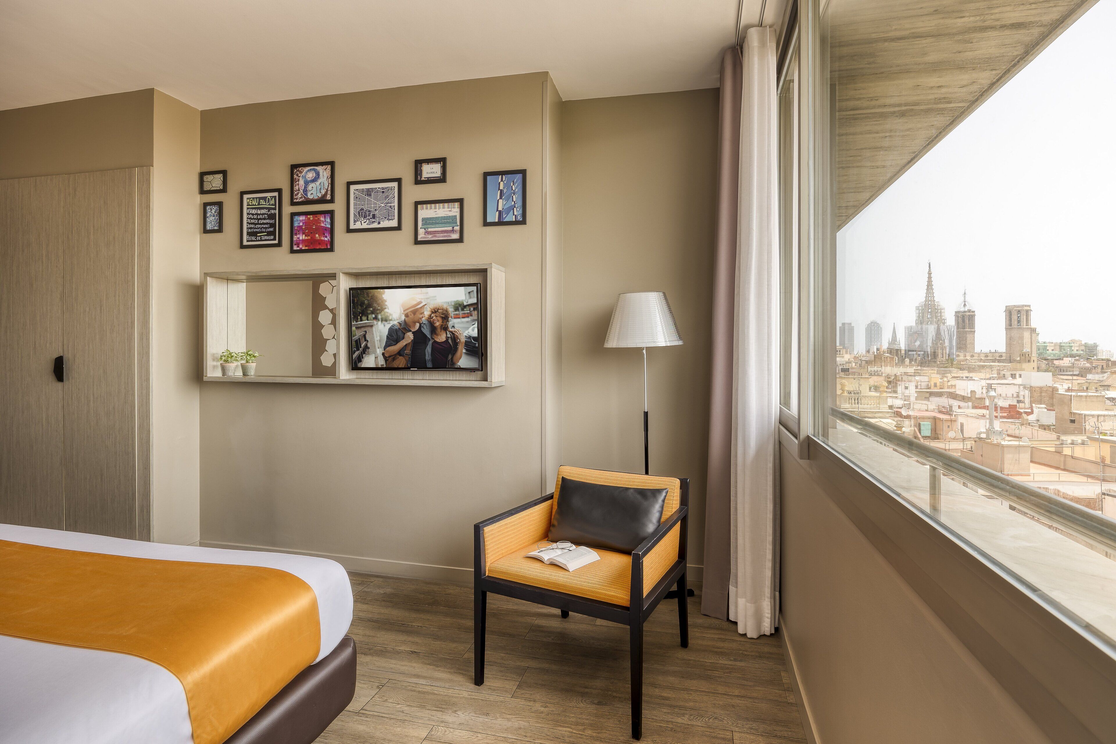 A hotel room with a bed, a chair, a city view, framed pictures on the wall, and a lamp.