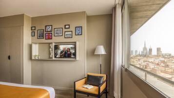 A hotel room with a bed, a chair, a city view, framed pictures on the wall, and a lamp.