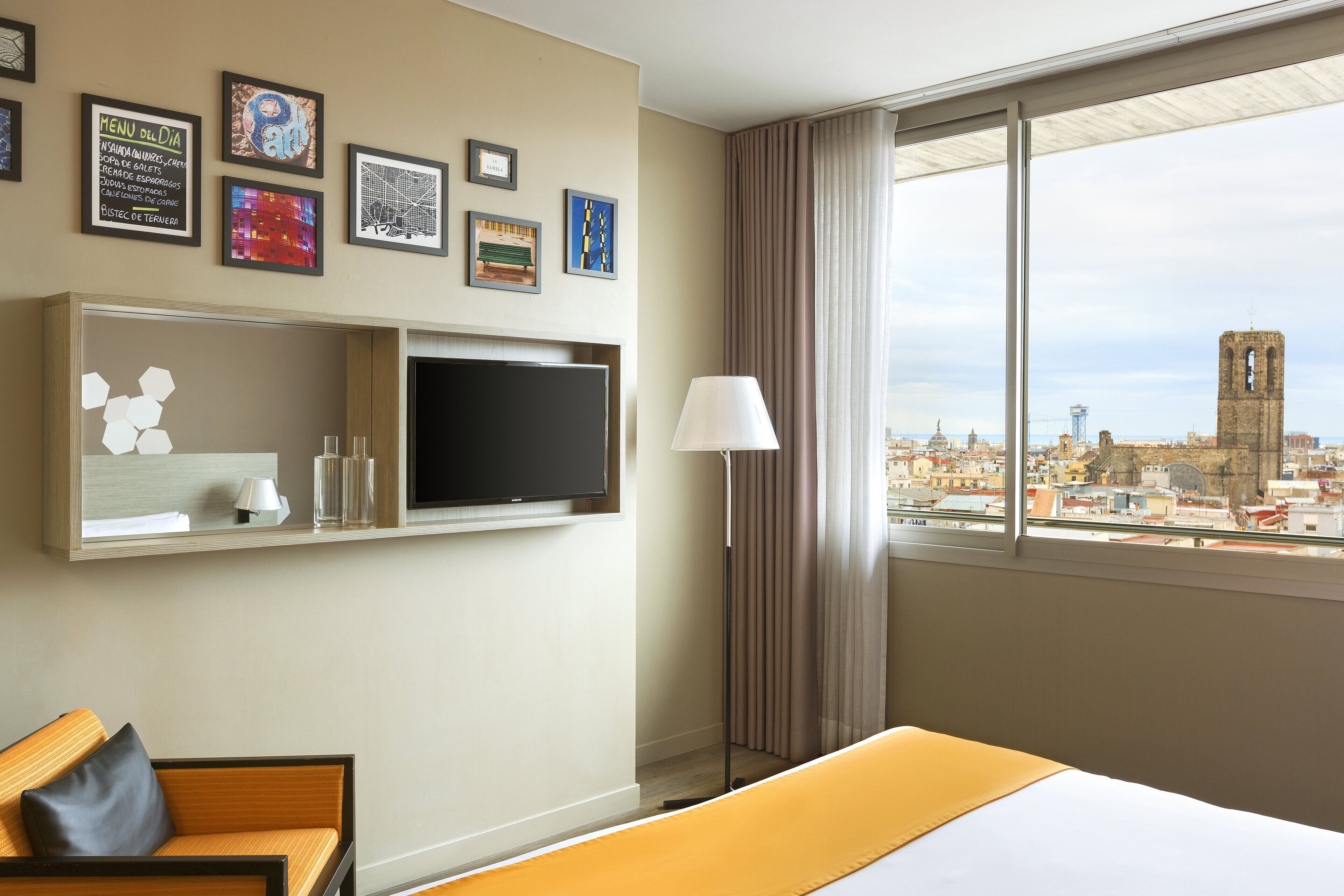 A hotel room with a bed, a TV mounted on the wall, a sofa, a lamp, and a city view through the window.