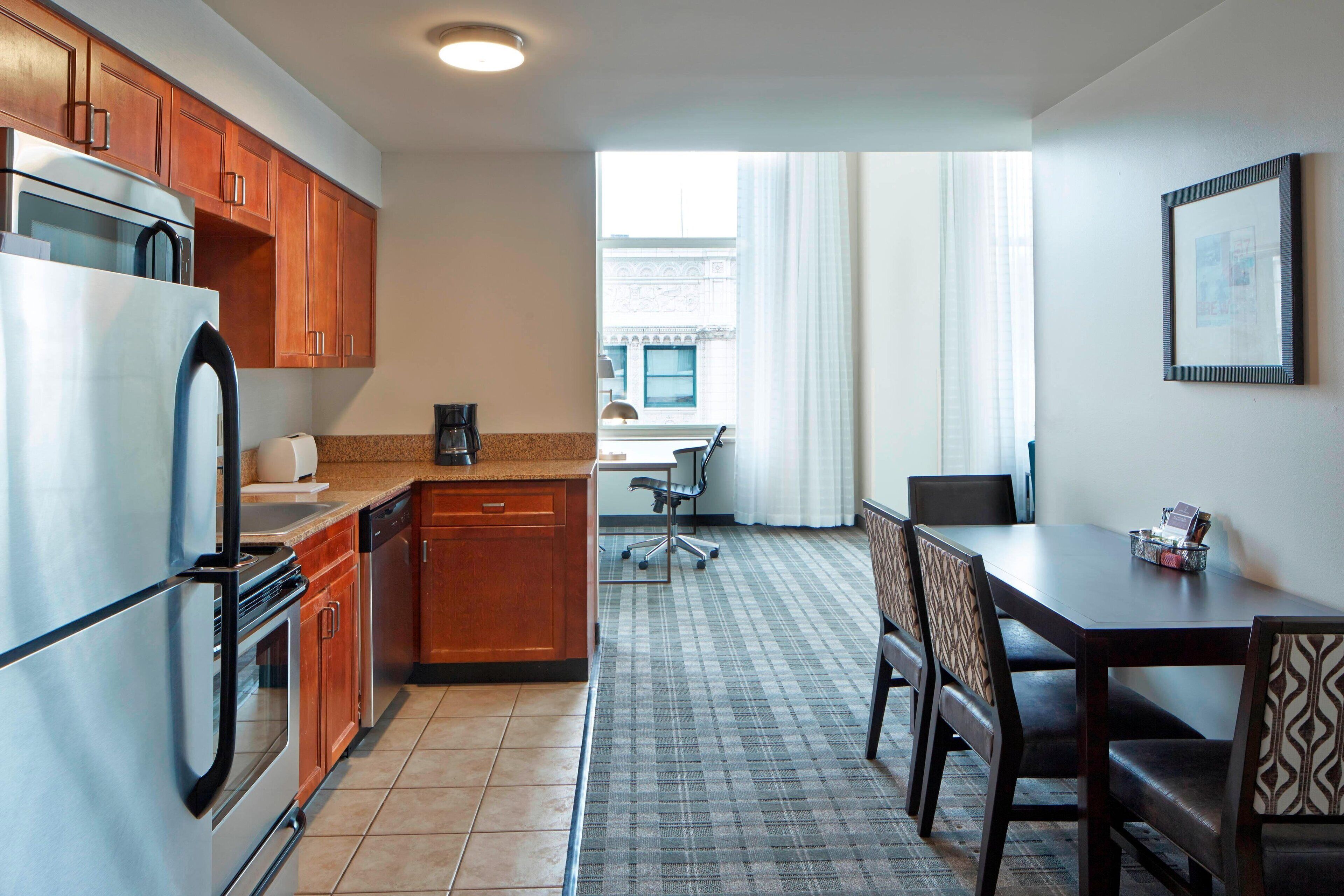 A compact kitchen with a refrigerator, stove, and wooden cabinets. There is a dining table with chairs and a window with curtains.