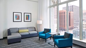 A modern hotel room with a gray sectional sofa, two blue armchairs, a small table, and a large window offering a city view. - Residence Inn Milwaukee Downtown by Marriott (Milwaukee)