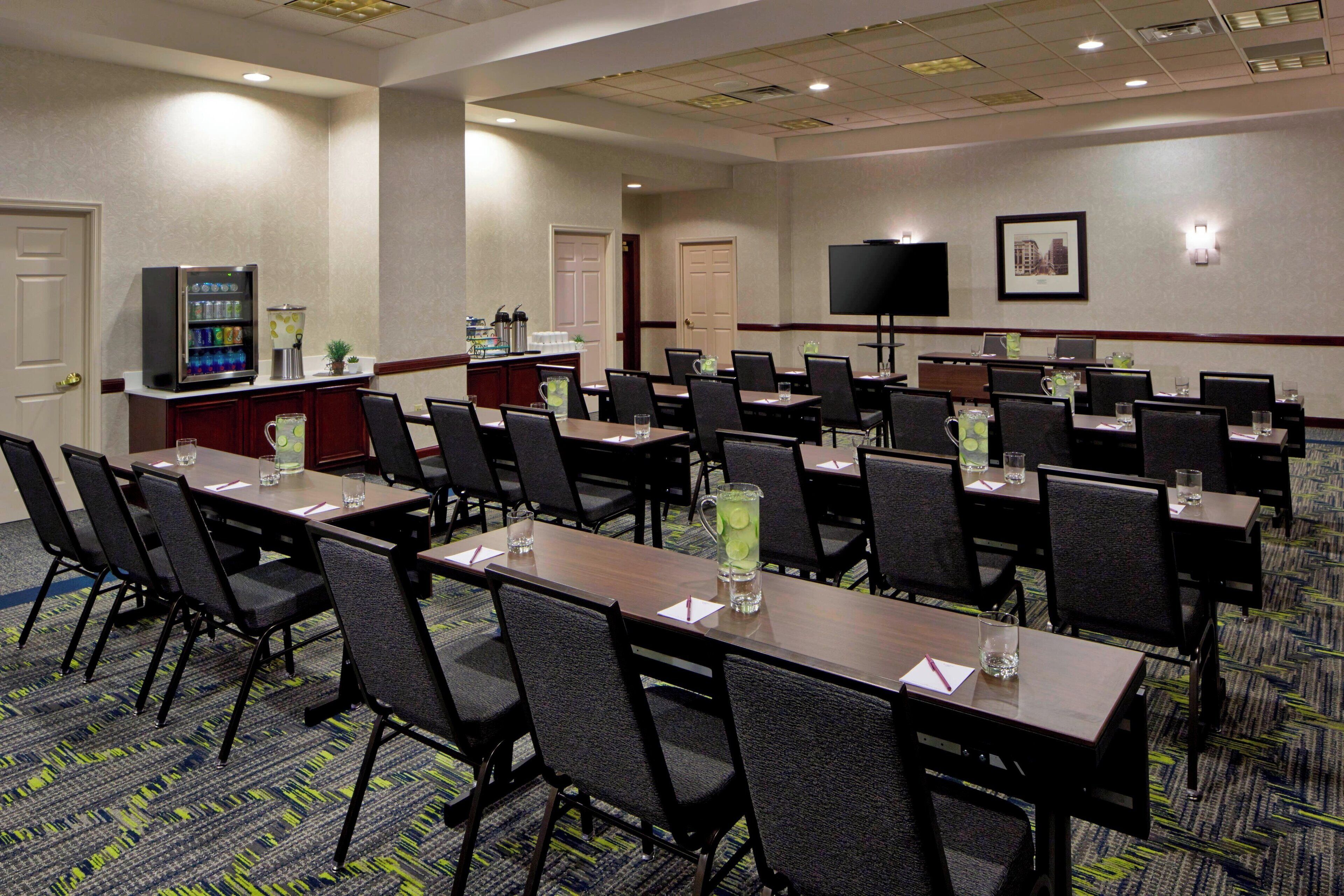 A conference room with multiple tables and chairs, a refrigerator with beverages, a television, and a small plant.
