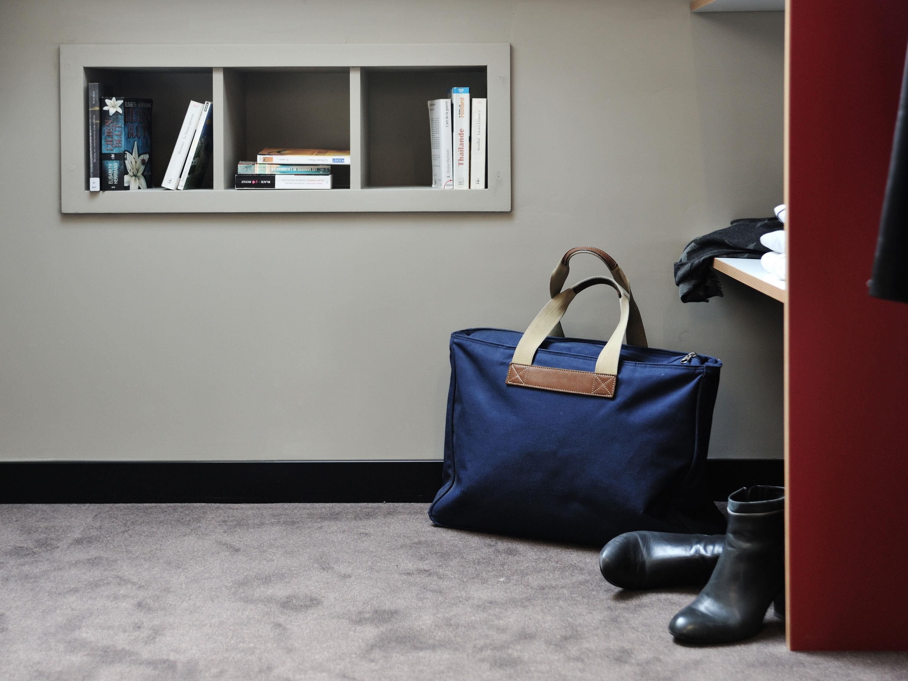 Une pièce avec un tapis gris, un sac bleu avec une bandoulière marron, des bottes noires et une bibliothèque contenant des livres et un tableau encadré.