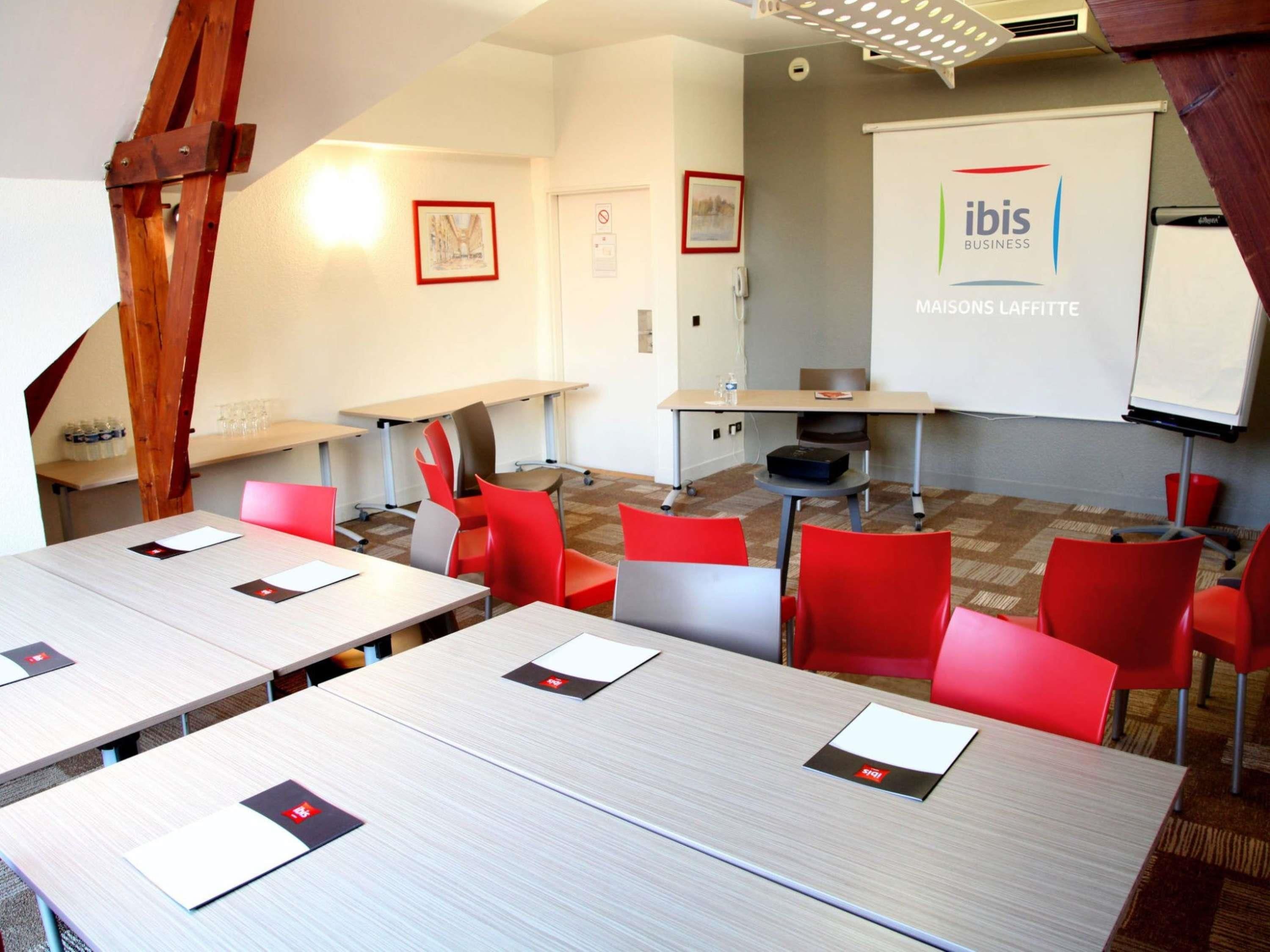 Una sala de conferencias con varias mesas y sillas, una pizarra y una pantalla proyectora que muestra 'ibis BUSINESS MAISONS LAFITTE'.