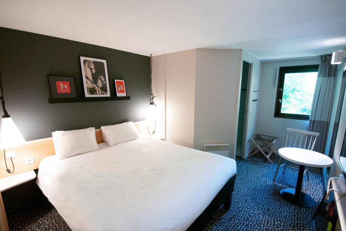 Habitación de hotel con una cama grande, una mesita pequeña y una silla. Hay cuadros en la pared y una ventana con cortinas.