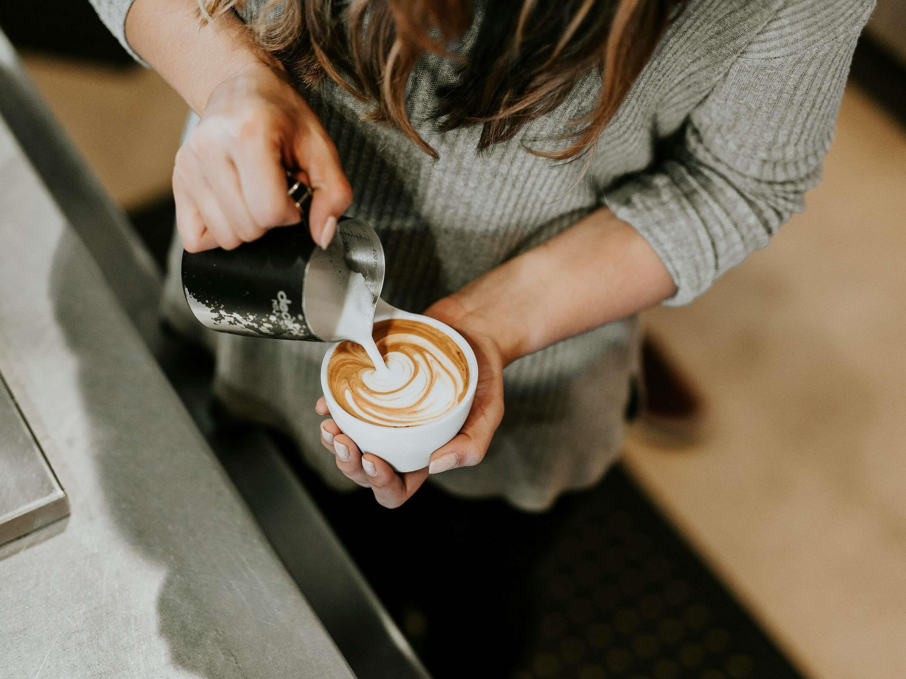 A person pouring milk into a cup of coffee, creating a latte art pattern.
