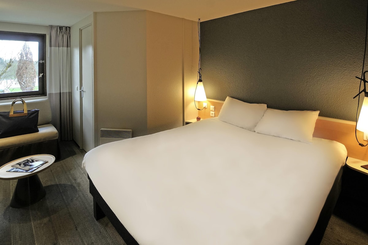 A hotel room with a large bed, bedside tables, a small table with a magazine, a window with curtains, and a view of trees outside.