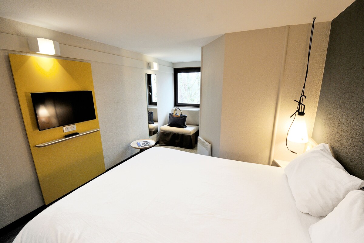 Une chambre d’hôtel avec un lit, un téléviseur fixé sur un panneau jaune, une petite table avec une chaise et une fenêtre donnant sur un paysage.