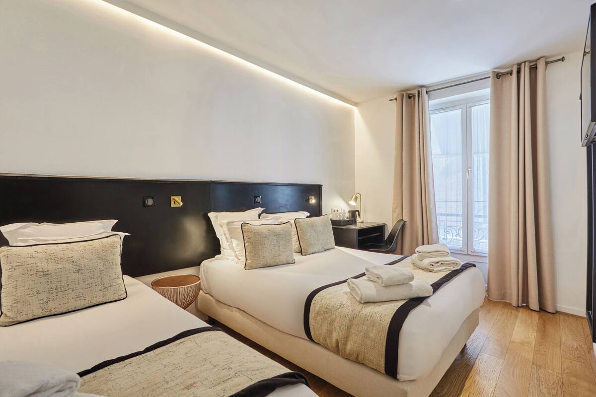 A hotel room with two beds, a desk, a chair, a window with curtains, and a flat-screen TV.