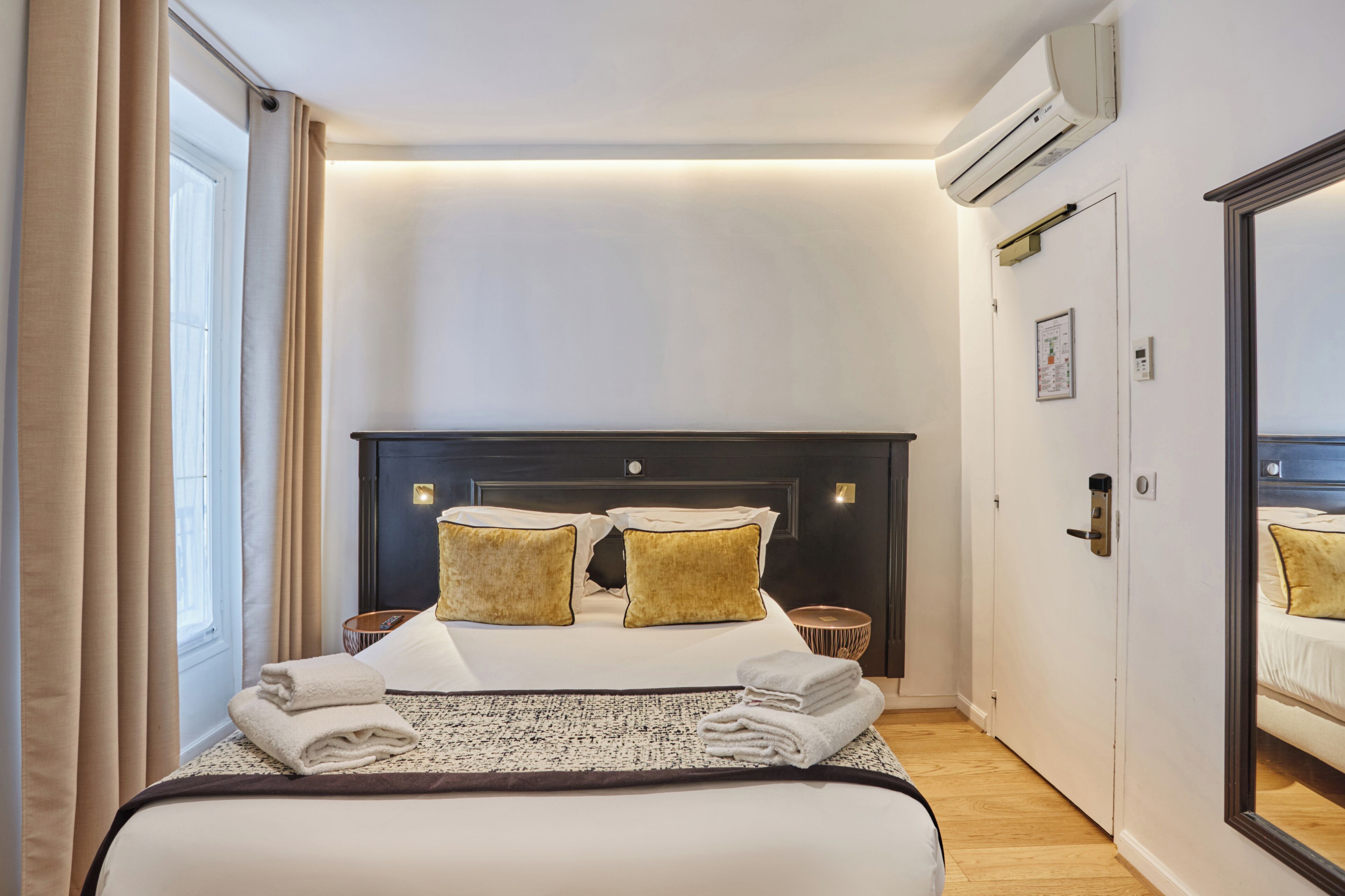 A hotel room with a bed, bedside tables, a mirror, and an air conditioning unit.