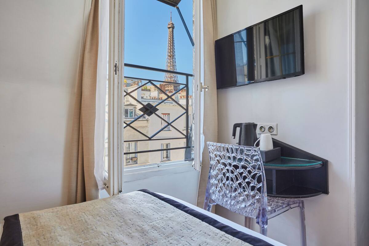 A hotel room with a bed, a desk with a chair, a TV mounted on the wall, and a view of the Eiffel Tower through a window.