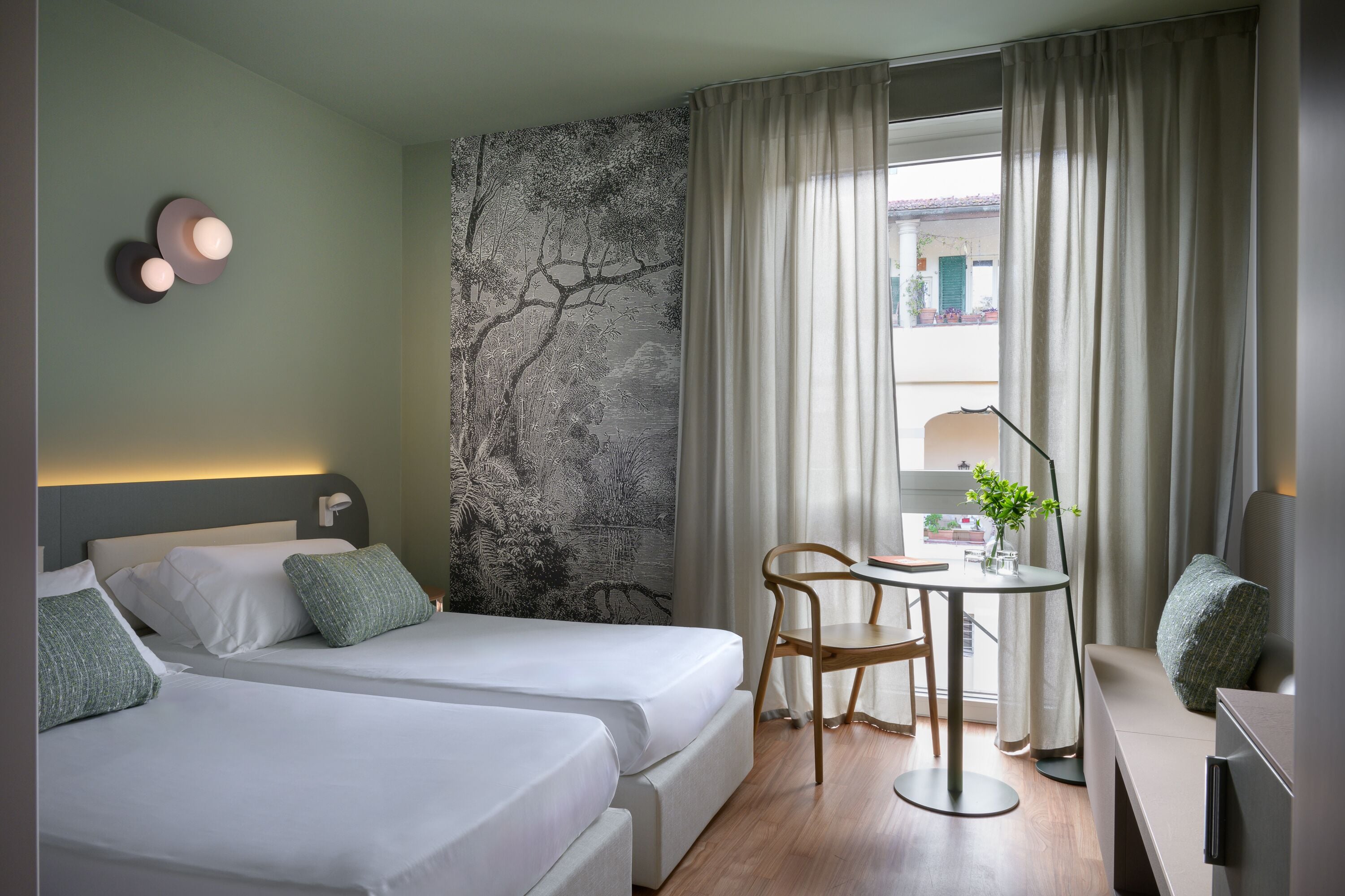 A hotel room with two beds, a desk with a chair, a lamp, and a view of the outdoors.
