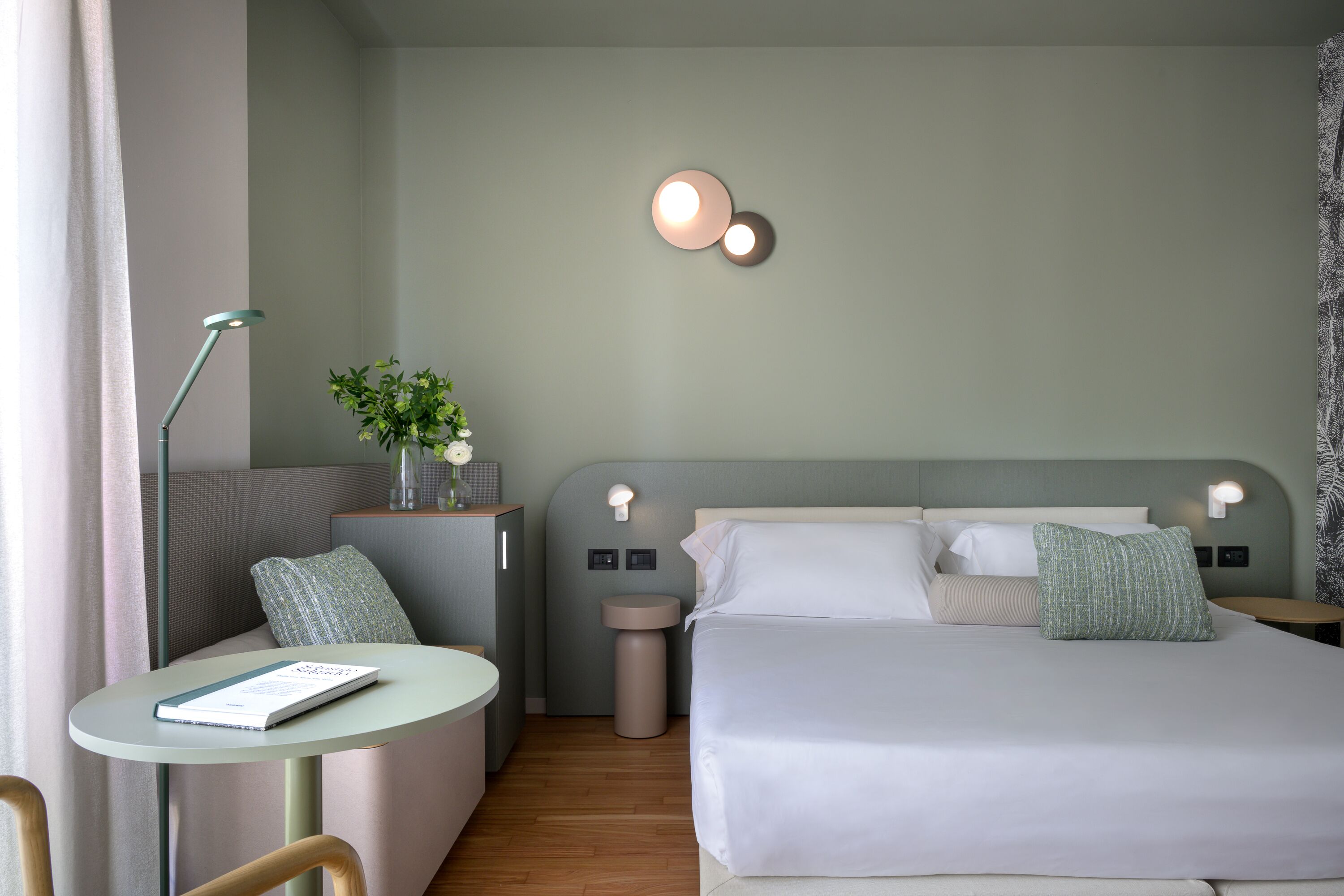 Habitación de hotel con cama, mesitas de noche, una pequeña mesa redonda, una silla y una lámpara.