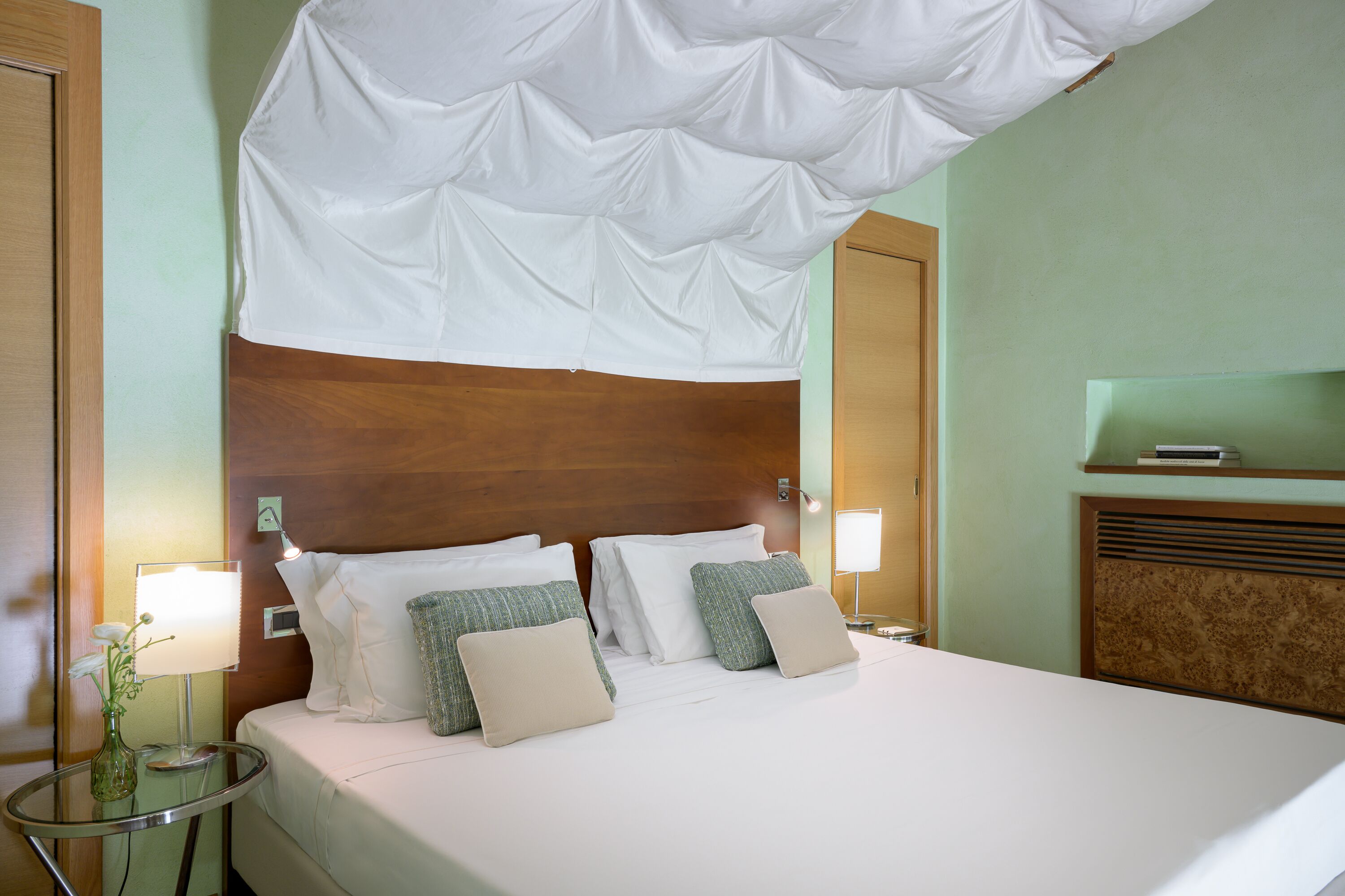 Una habitación de hotel con una cama grande, dos mesitas de noche, una lámpara, un florero con flores y un cabecero de madera.
