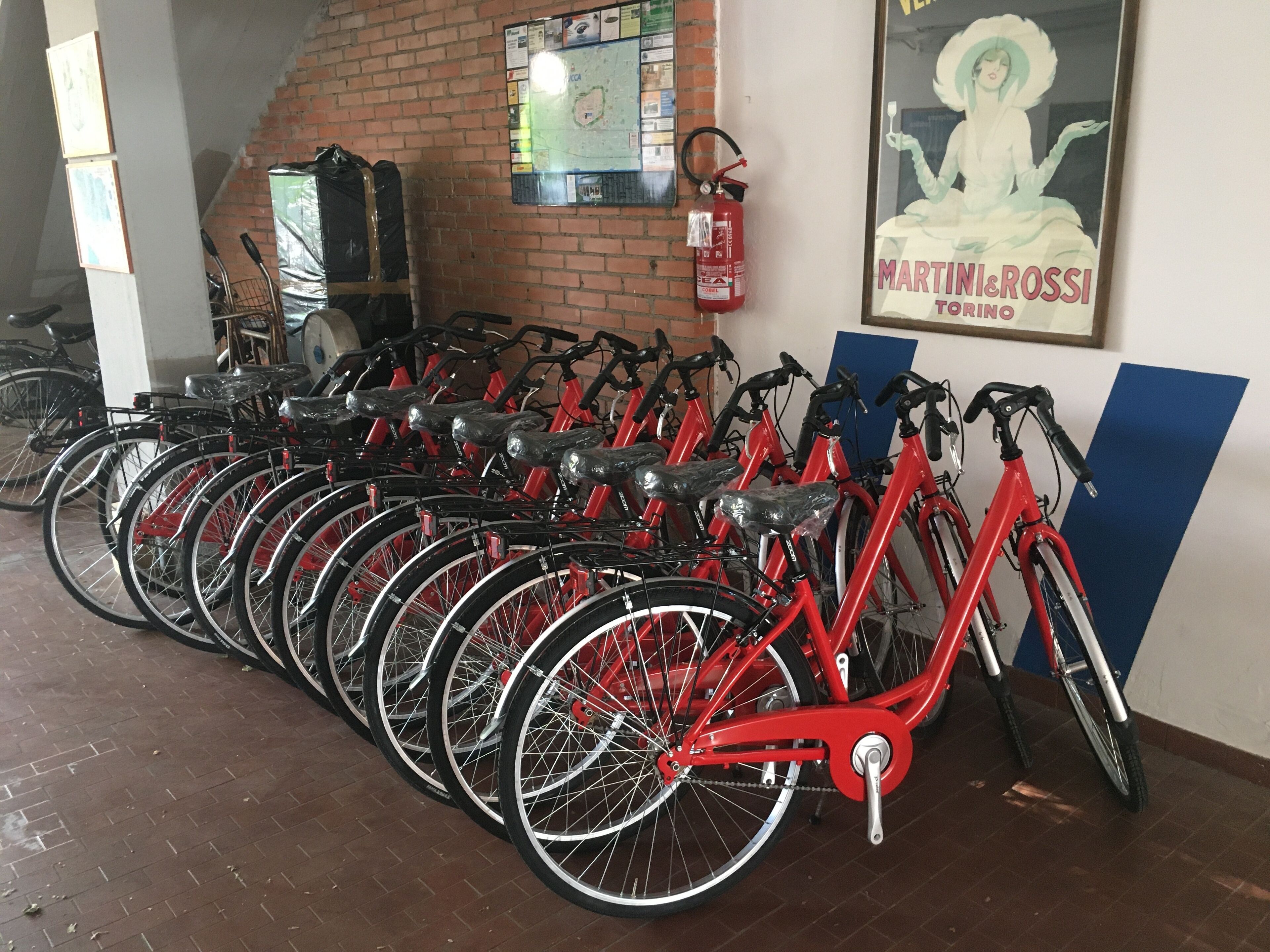 Een rij rode fietsen geparkeerd binnen, een brandblusser aan de muur en een poster van een vrouw.
