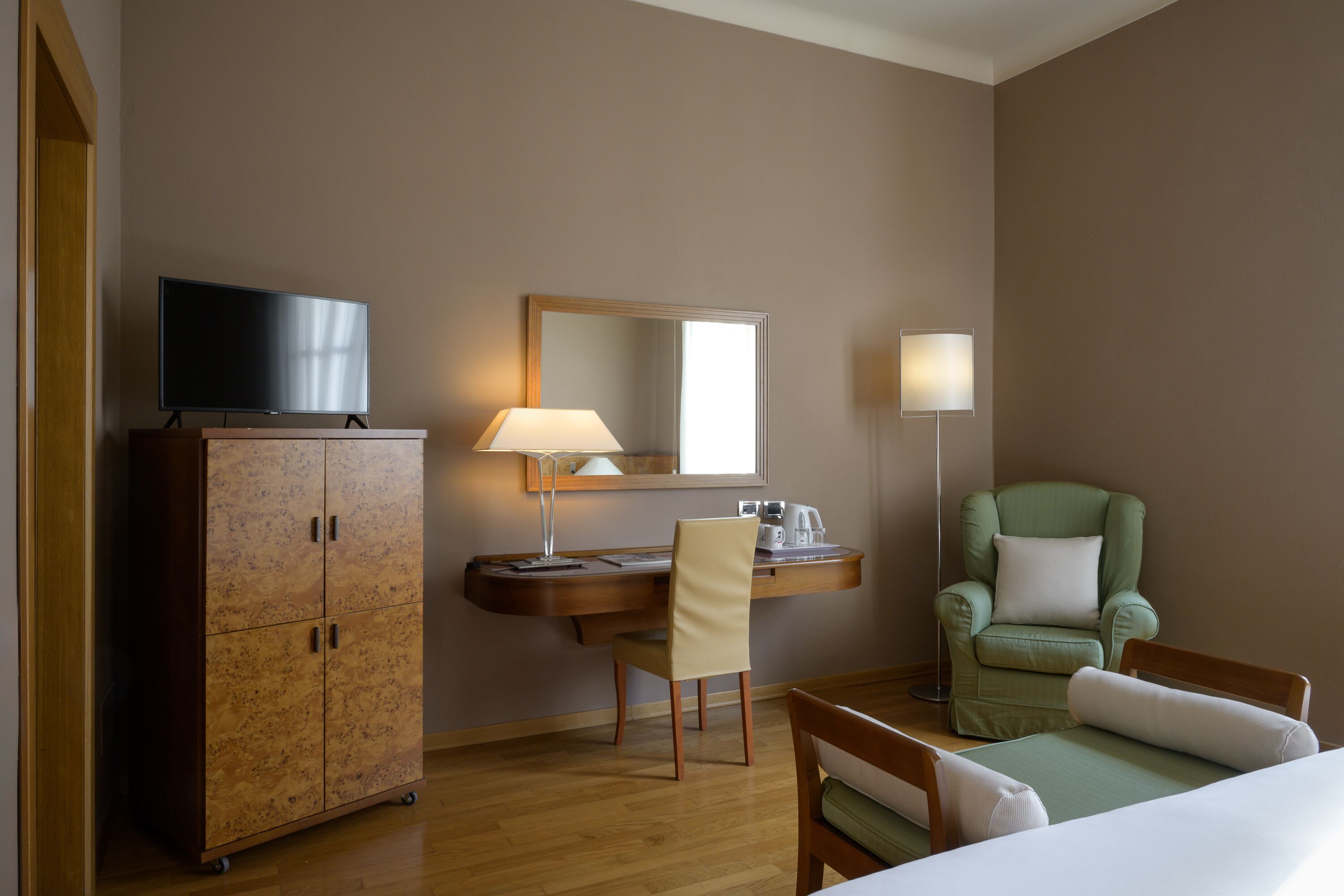 Quarto de hotel com guarda-roupa de madeira, escrivaninha com luminária, poltrona verde e cama.