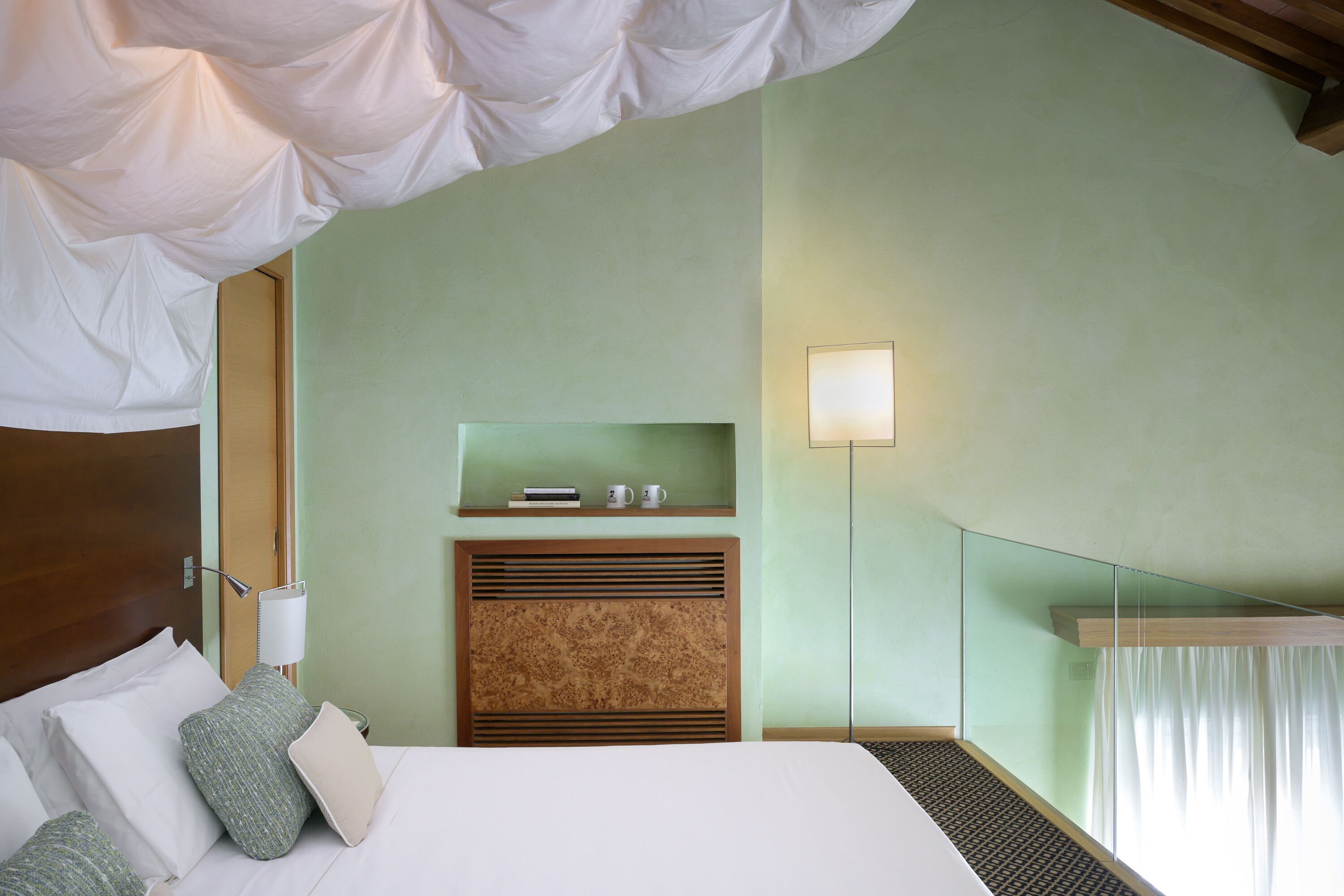 A hotel room with a bed, pillows, a nightstand, a lamp, a shelf with books and cups, and a window with curtains.