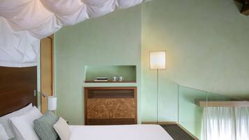A hotel room with a bed, pillows, a nightstand, a lamp, a shelf with books and cups, and a window with curtains.