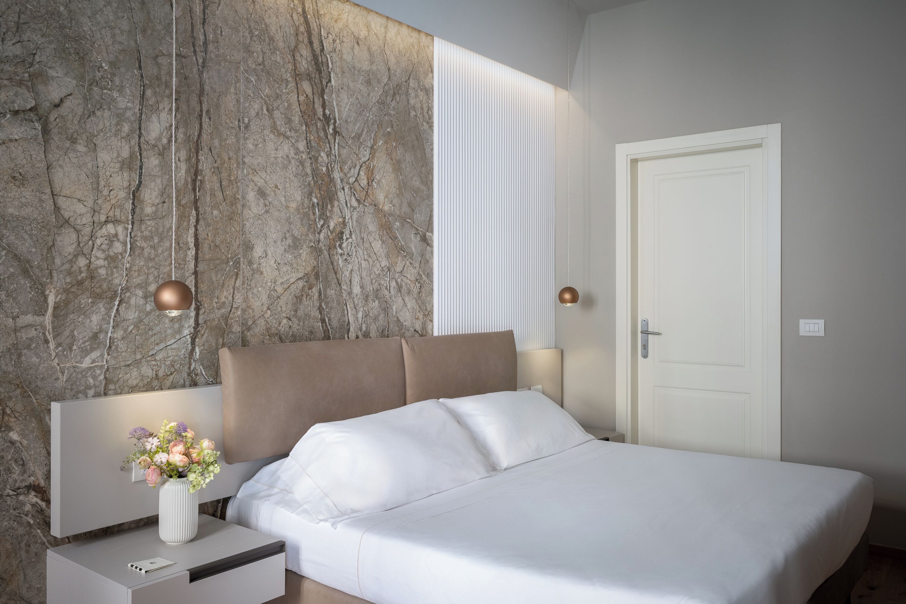 Ein modernes Schlafzimmer mit Marmorwand, einem Bett mit weißer Bettwäsche, einem Nachttisch mit Blumenvase und einer Tür mit Lichtschalter.