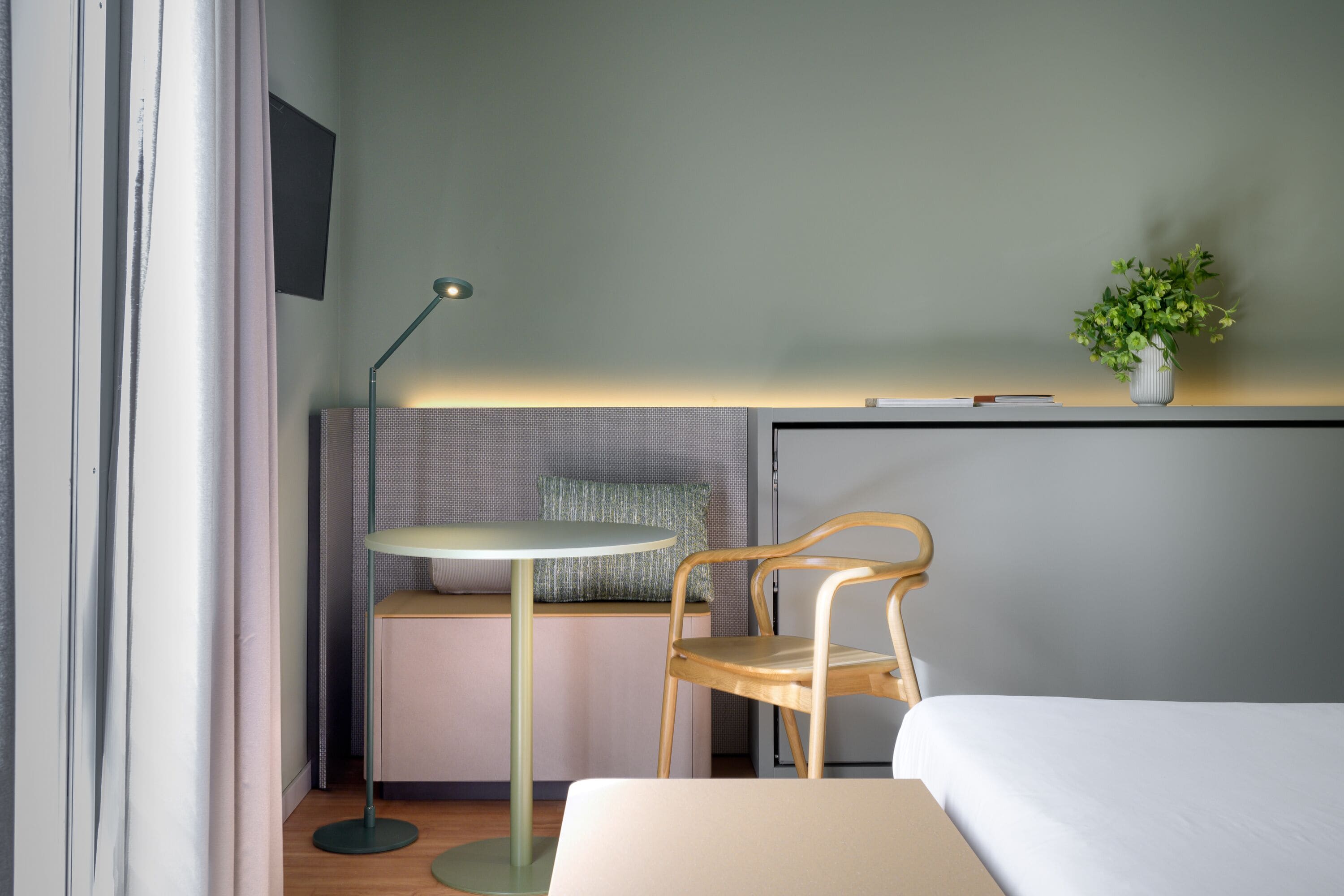 A hotel room with a bed, a desk with a chair, a standing lamp, a small table, and a potted plant.