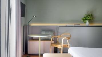 A hotel room with a bed, a desk with a chair, a standing lamp, a small table, and a potted plant.