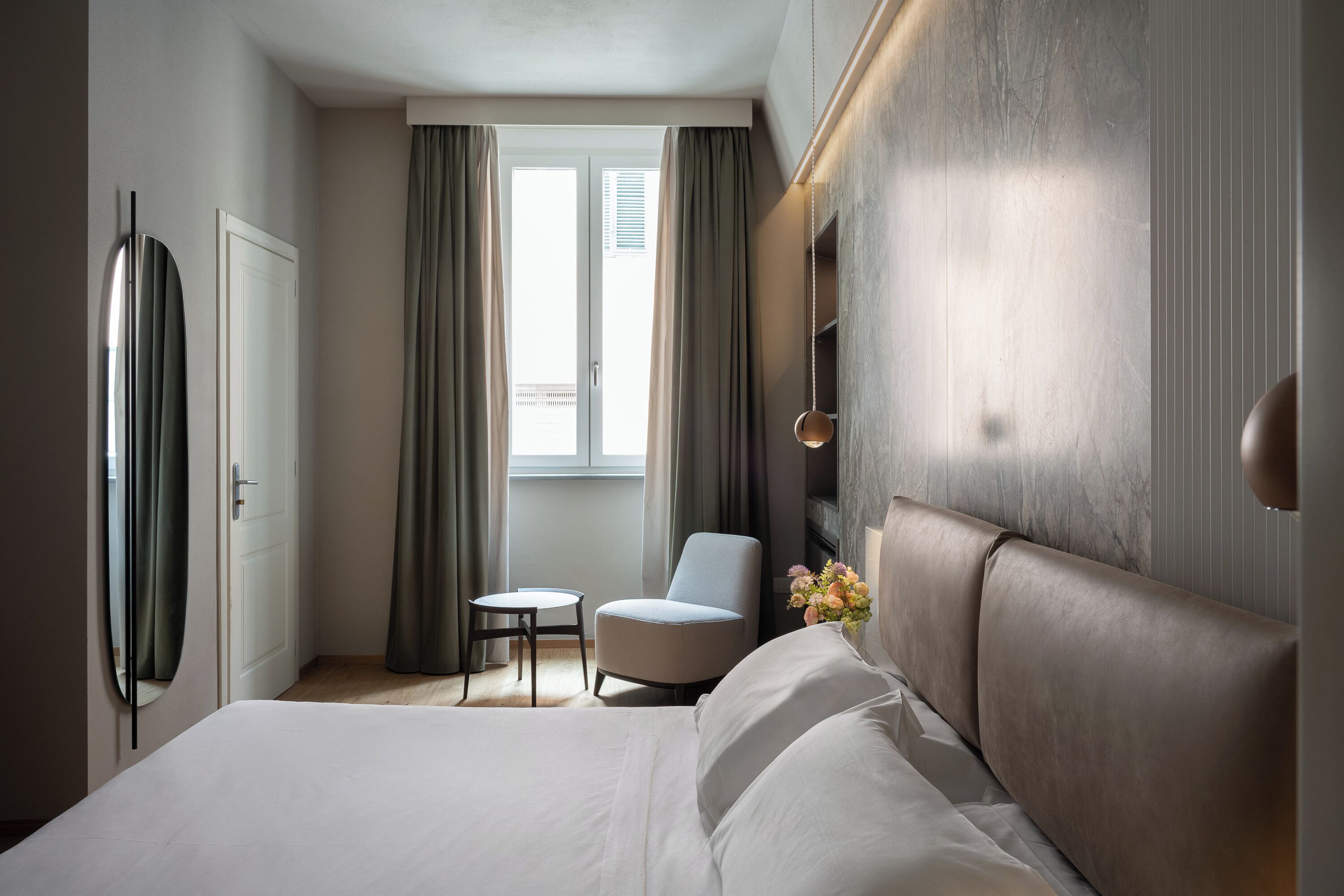 Une chambre d’hôtel moderne avec un grand lit, une petite table ronde, un fauteuil confortable et une vue sur l’extérieur grâce à une fenêtre avec des rideaux.