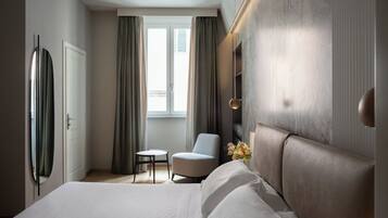 Une chambre d’hôtel moderne avec un grand lit, une petite table ronde, un fauteuil confortable et une vue sur l’extérieur grâce à une fenêtre avec des rideaux.