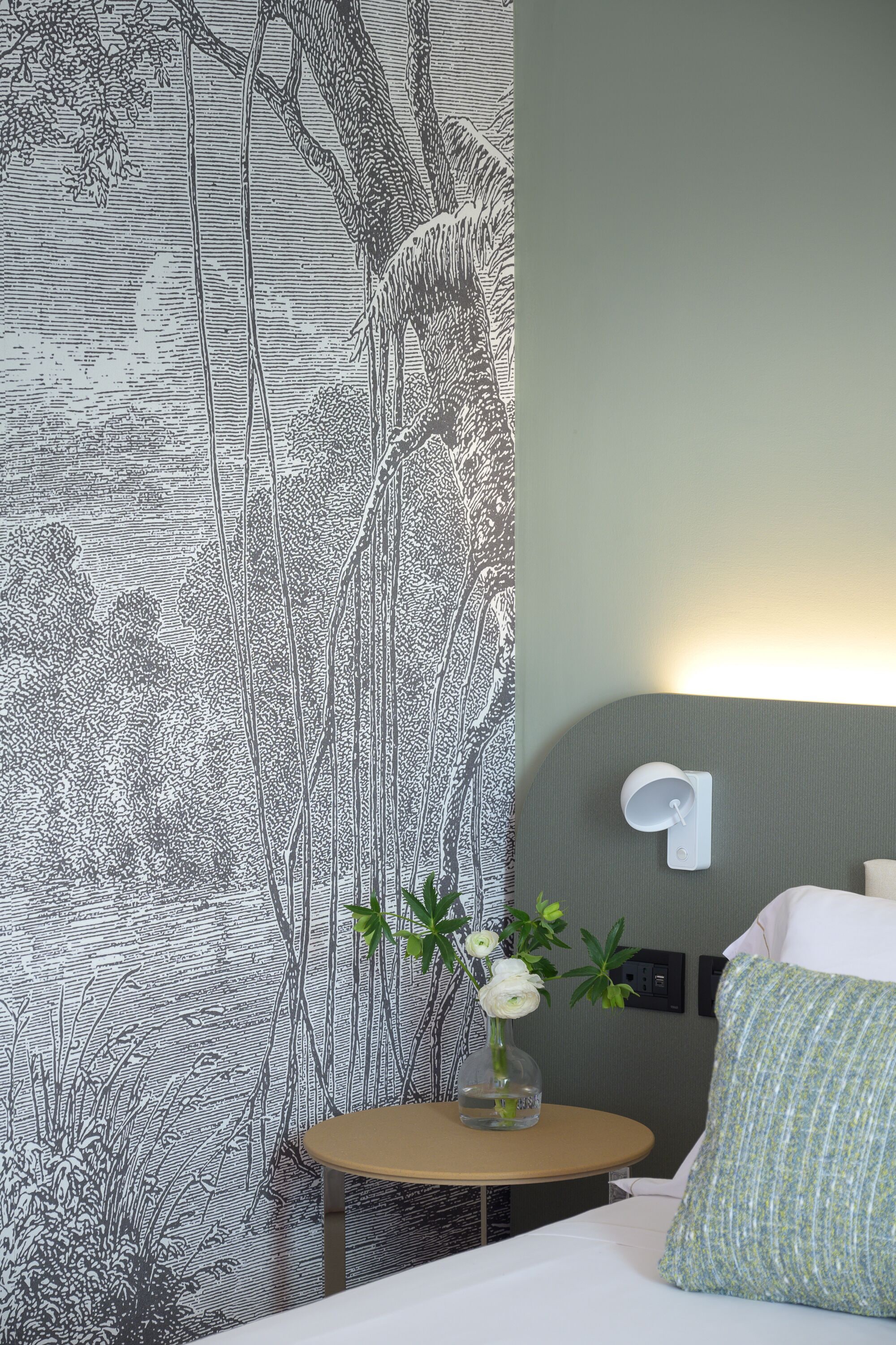 Un dormitorio con un amplio papel tapiz estampado, una mesita de noche con un jarrón de flores y un cabecero moderno.