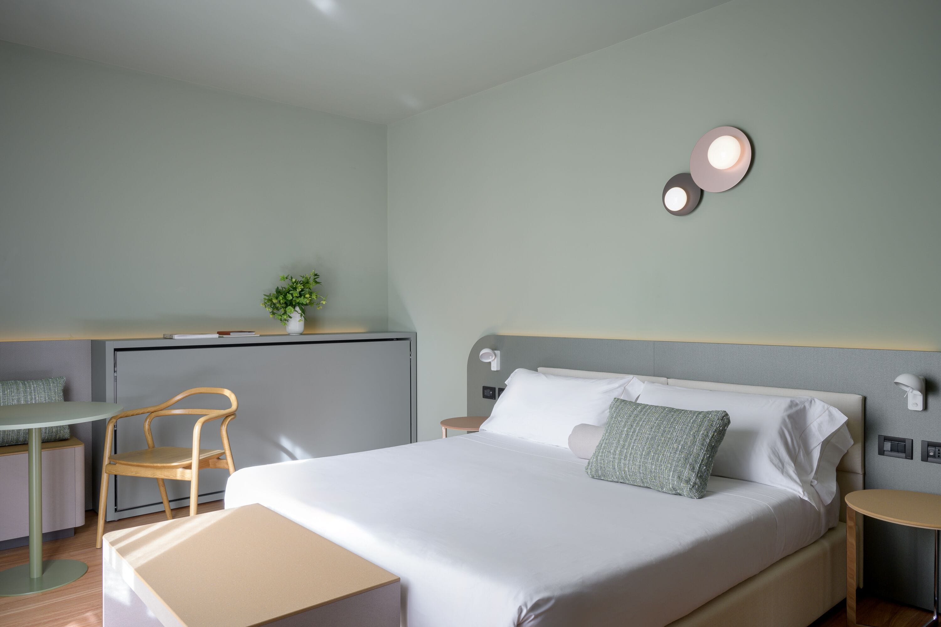 A modern hotel room with a bed, a desk, a chair, and a small table.
