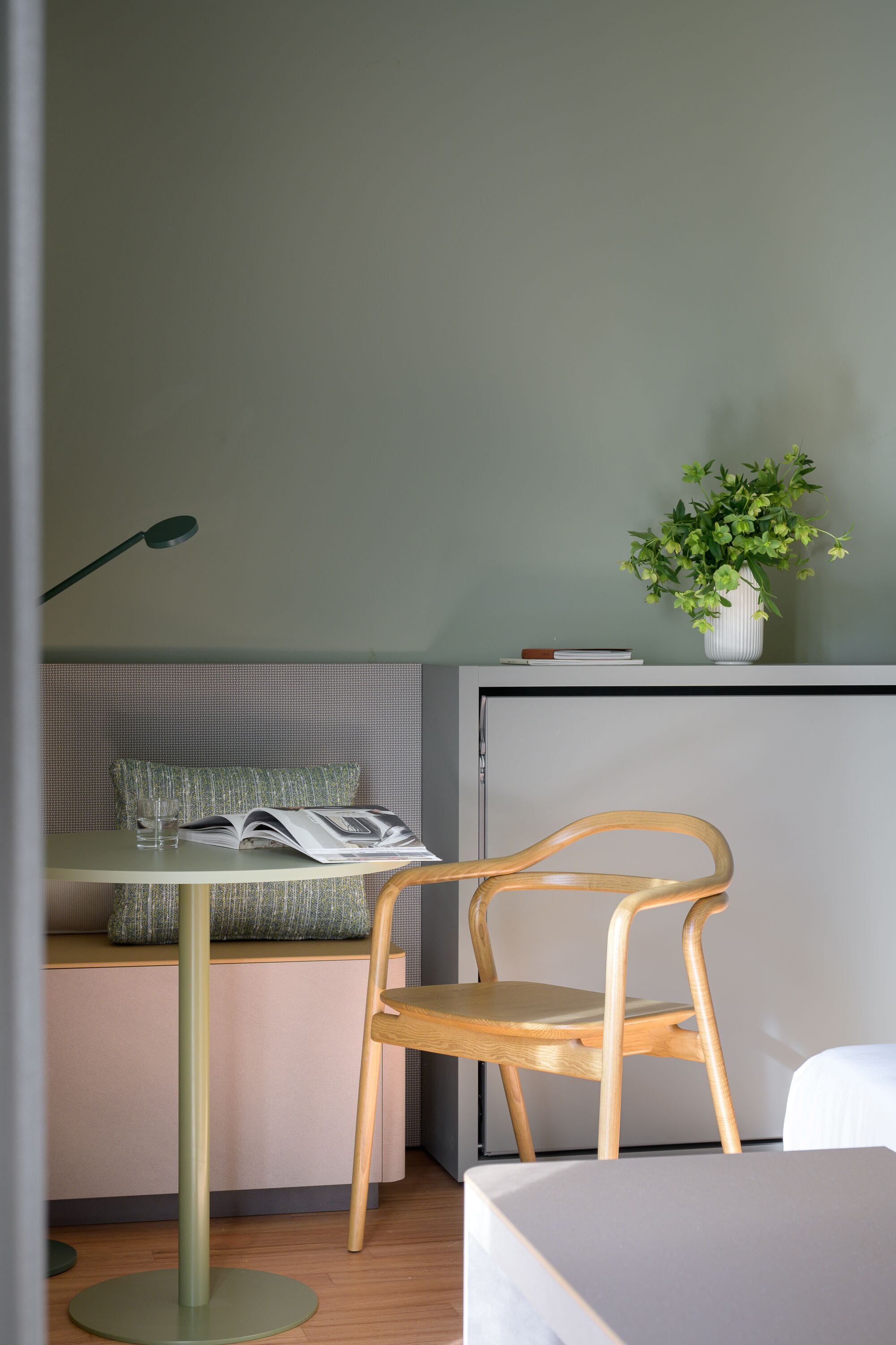 A room with a wooden chair, a small table with a book and glass, a potted plant, and a bed.
