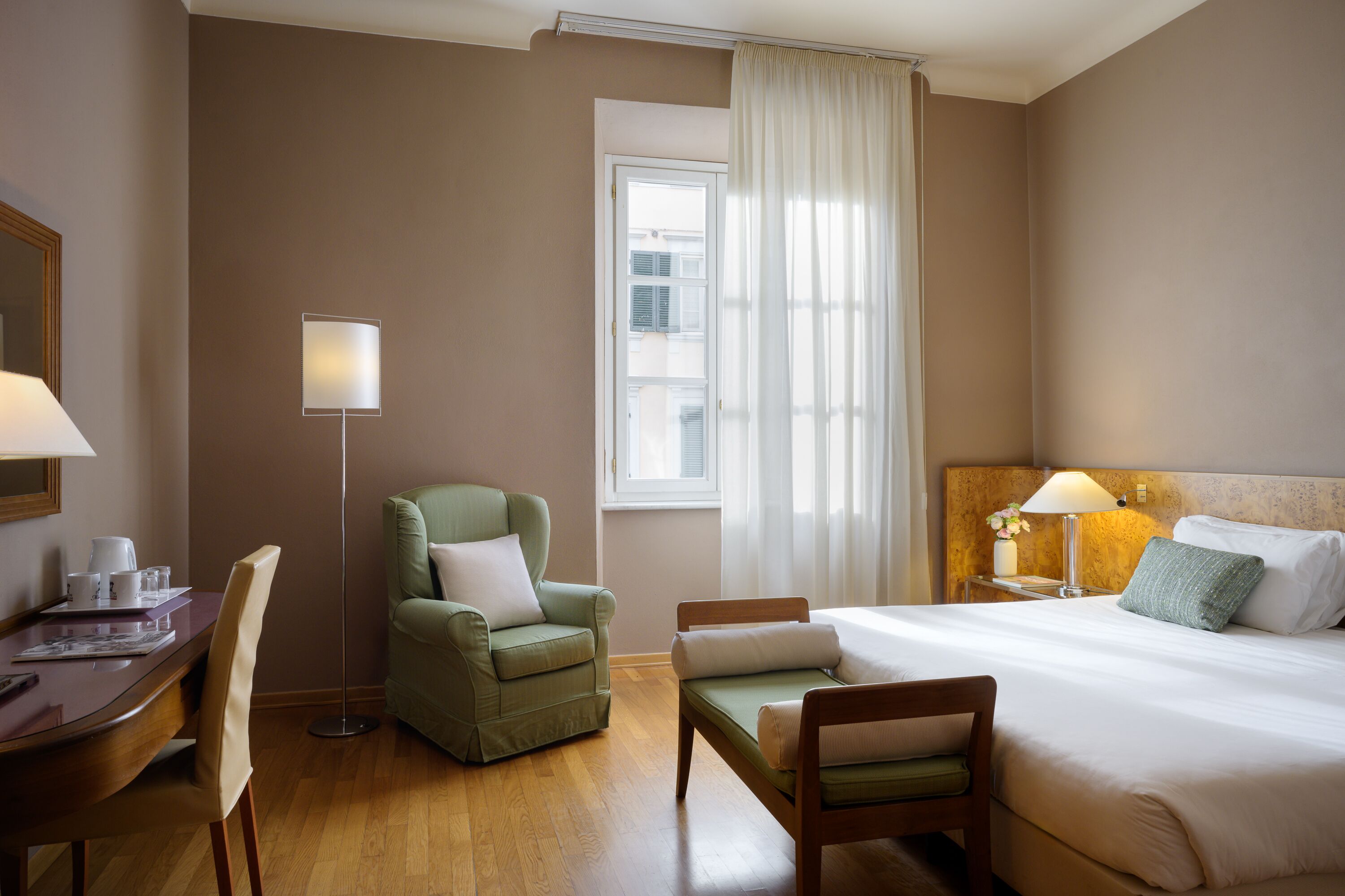 A hotel room with a bed, two chairs, a desk, a lamp, and a window with curtains.