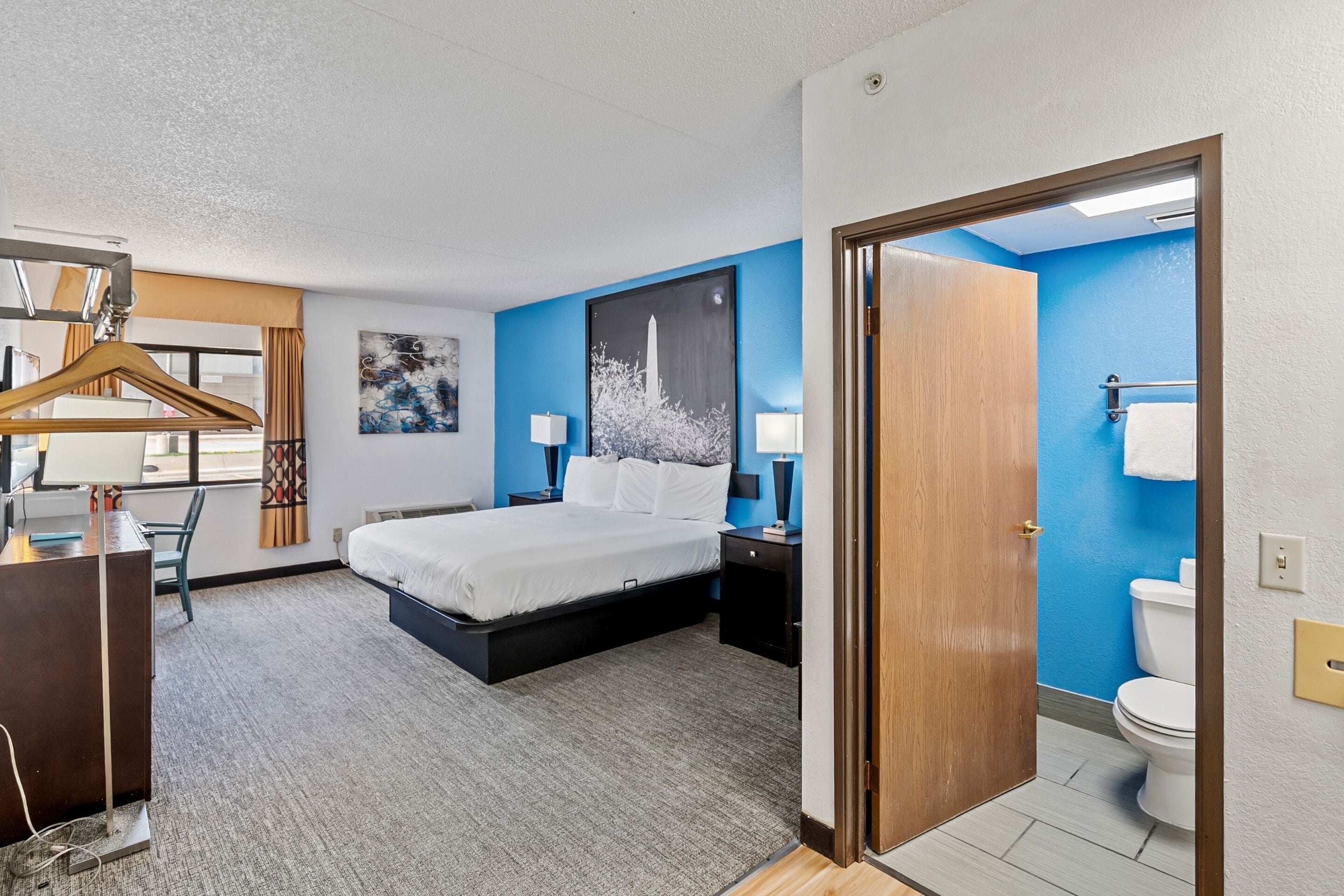 A hotel room with a bed, bedside tables, a desk, a chair, a hanging coat rack, a painting, and a bathroom with a toilet and towel rack.