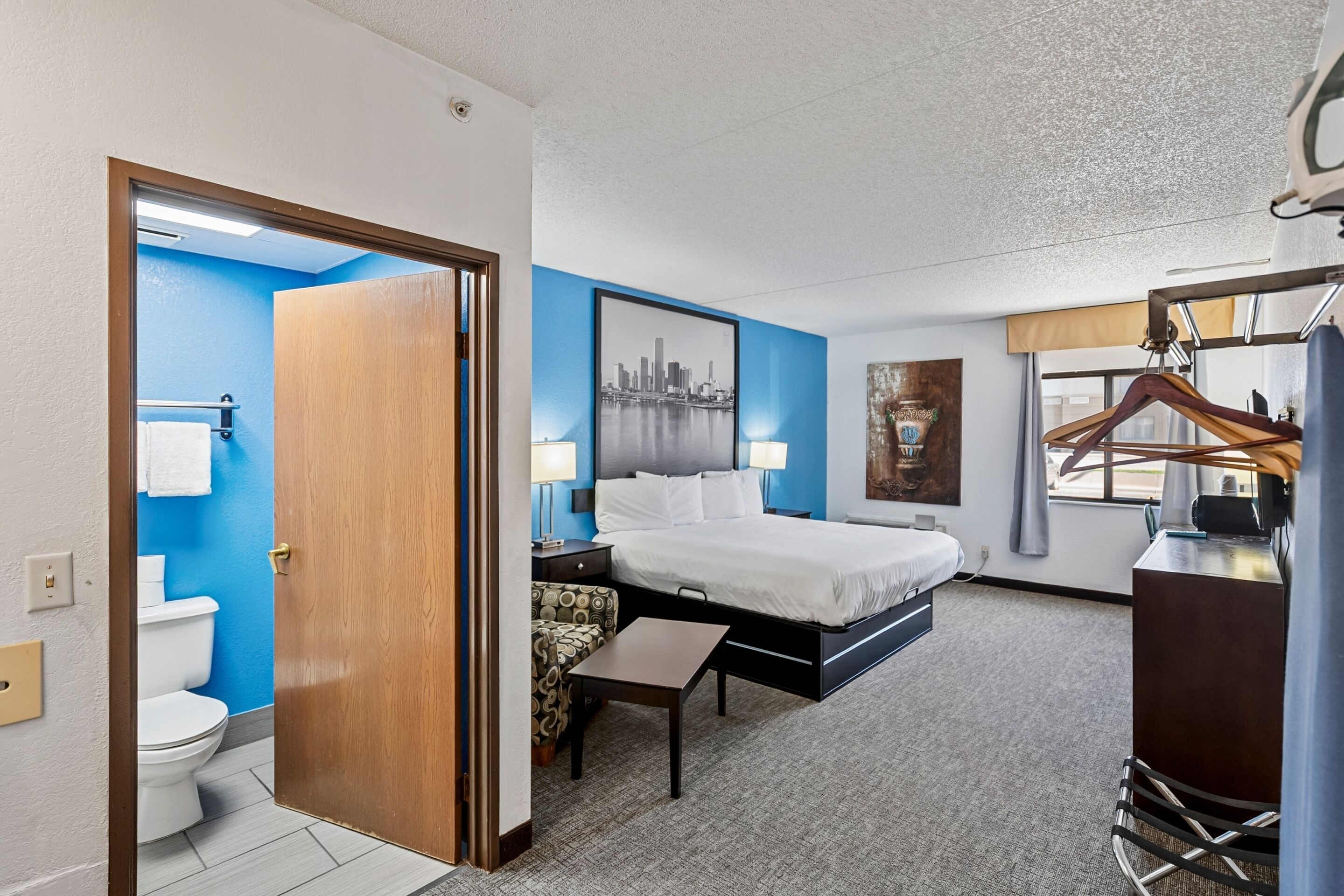 Habitación de hotel con cama, escritorio, silla, perchero y baño con inodoro y lavamanos.