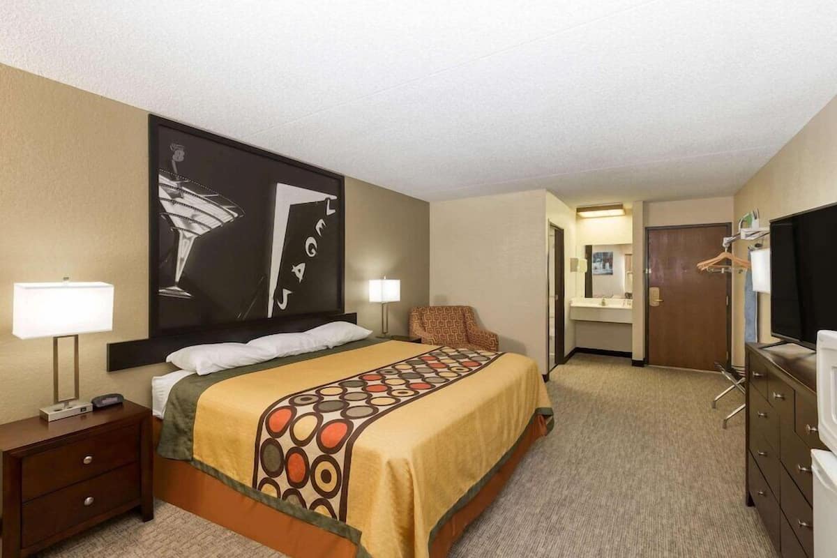 A hotel room with a bed, bedside lamps, a dresser, a television, a door leading to a bathroom, and a large abstract painting on the wall.