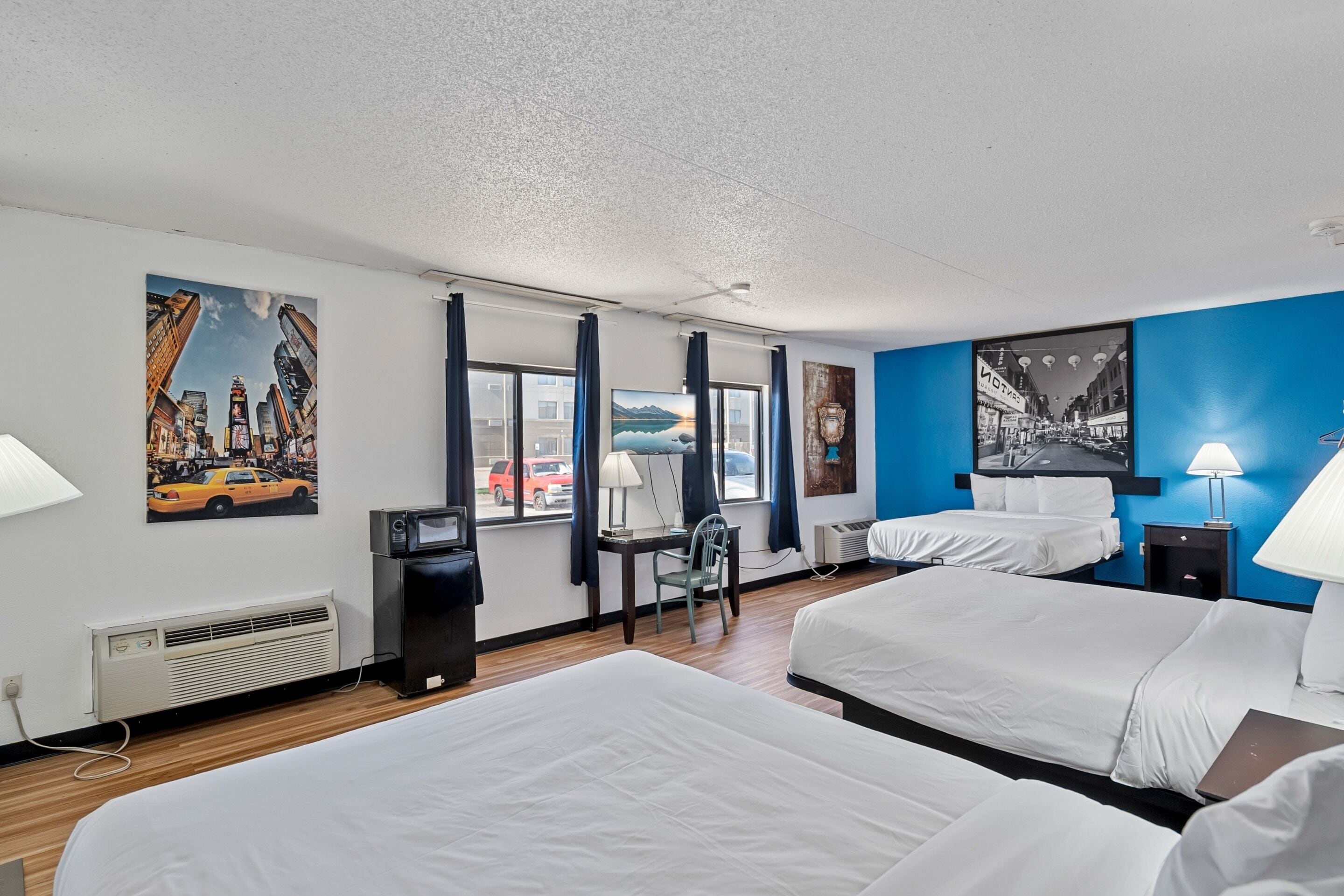 A hotel room with two beds, a desk, a chair, a window with curtains, and wall art.