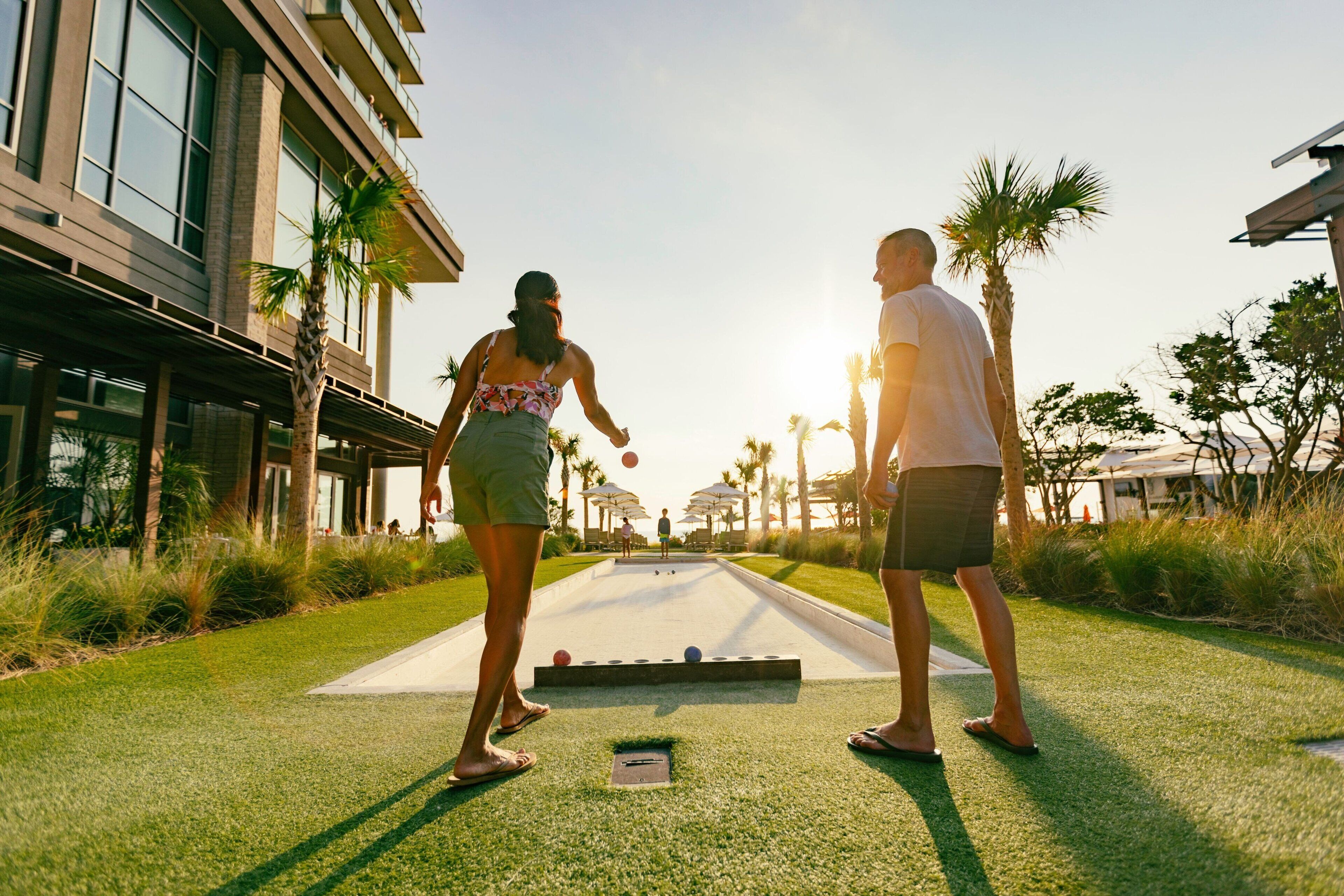 A courtyard with a shuffleboard table, palm trees, and a building with large windows.