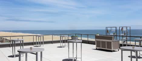 A rooftop terrace with tables and chairs, overlooking a beach and the ocean.