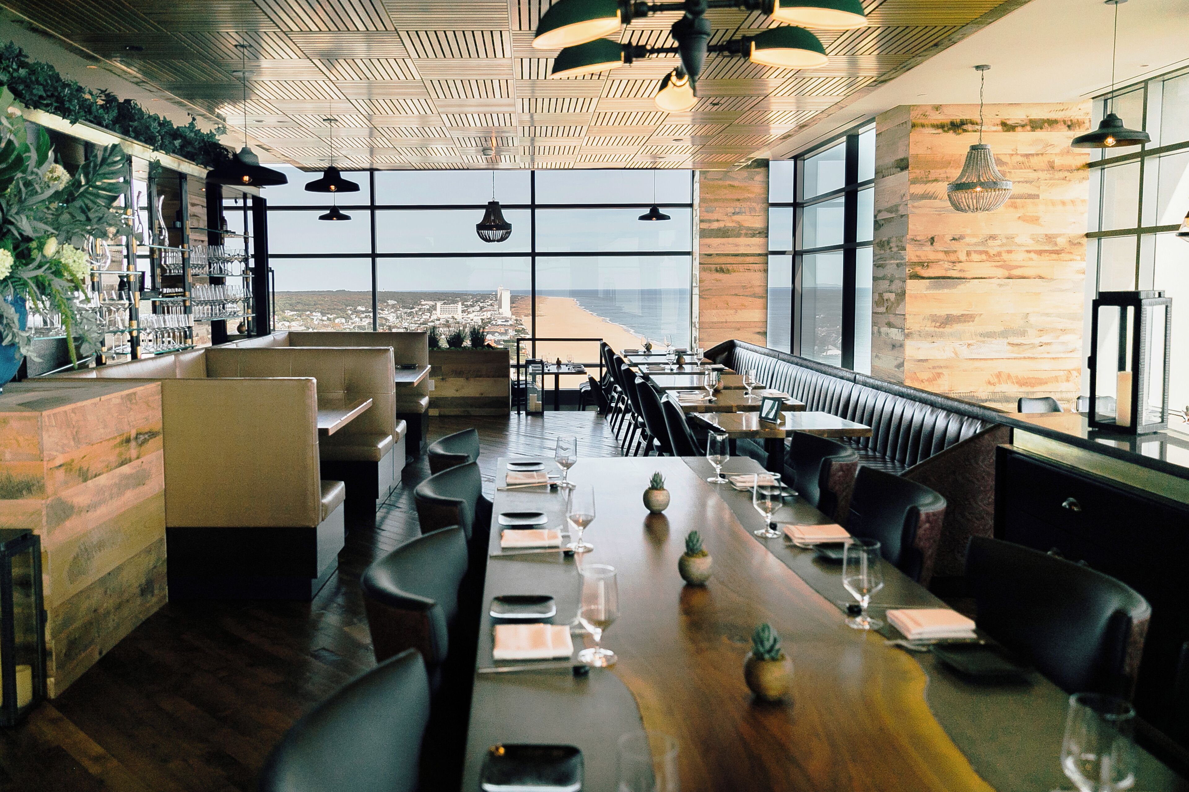 Un restaurante con una larga mesa de madera dispuesta para comer, amplias ventanas que ofrecen vistas a la ciudad y una zona de bar con plantas y vasos.