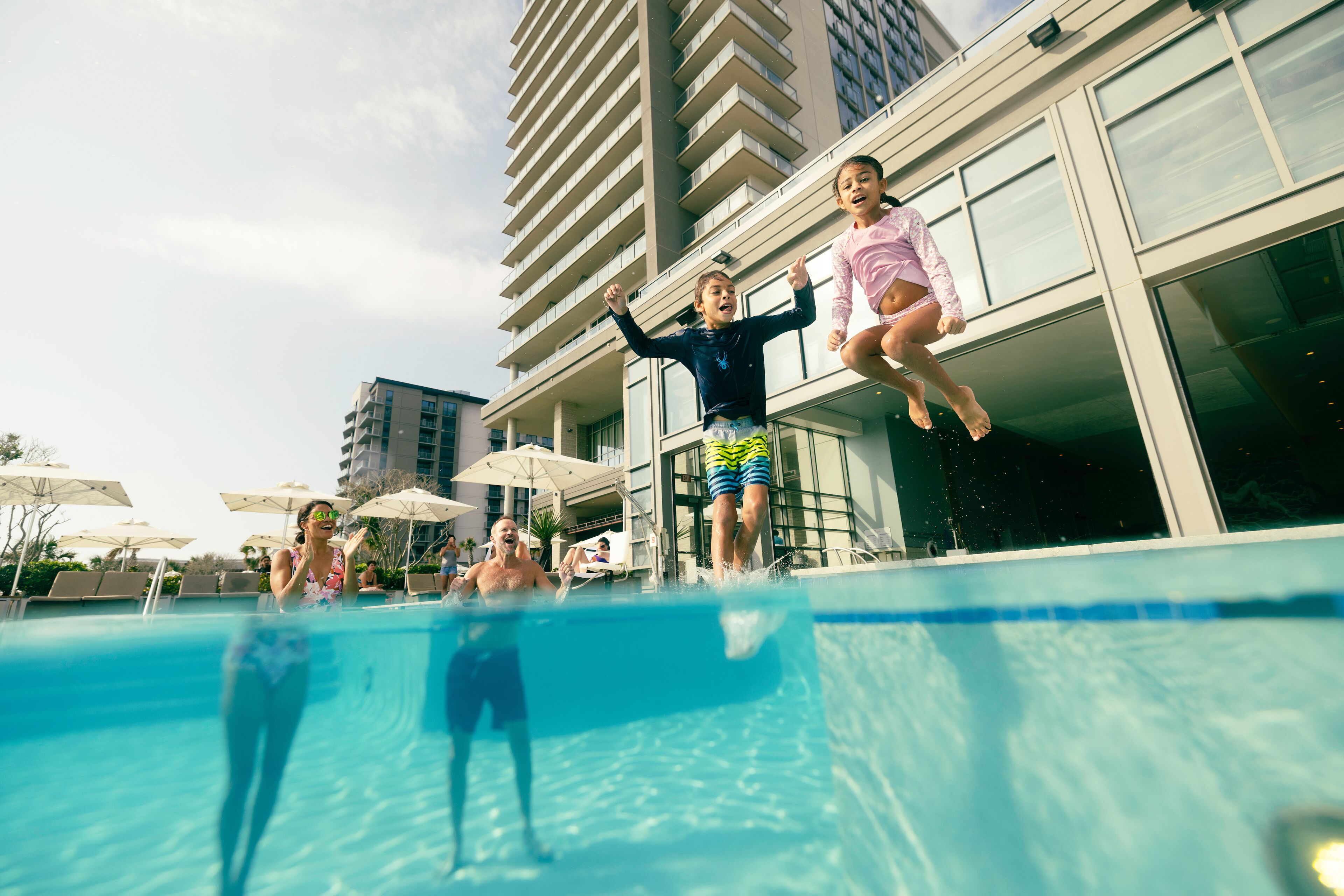 Una piscina con gente saltando, rodeada de edificios altos y sombrillas.