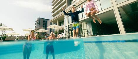 A swimming pool with people jumping into it, surrounded by tall buildings and umbrellas.