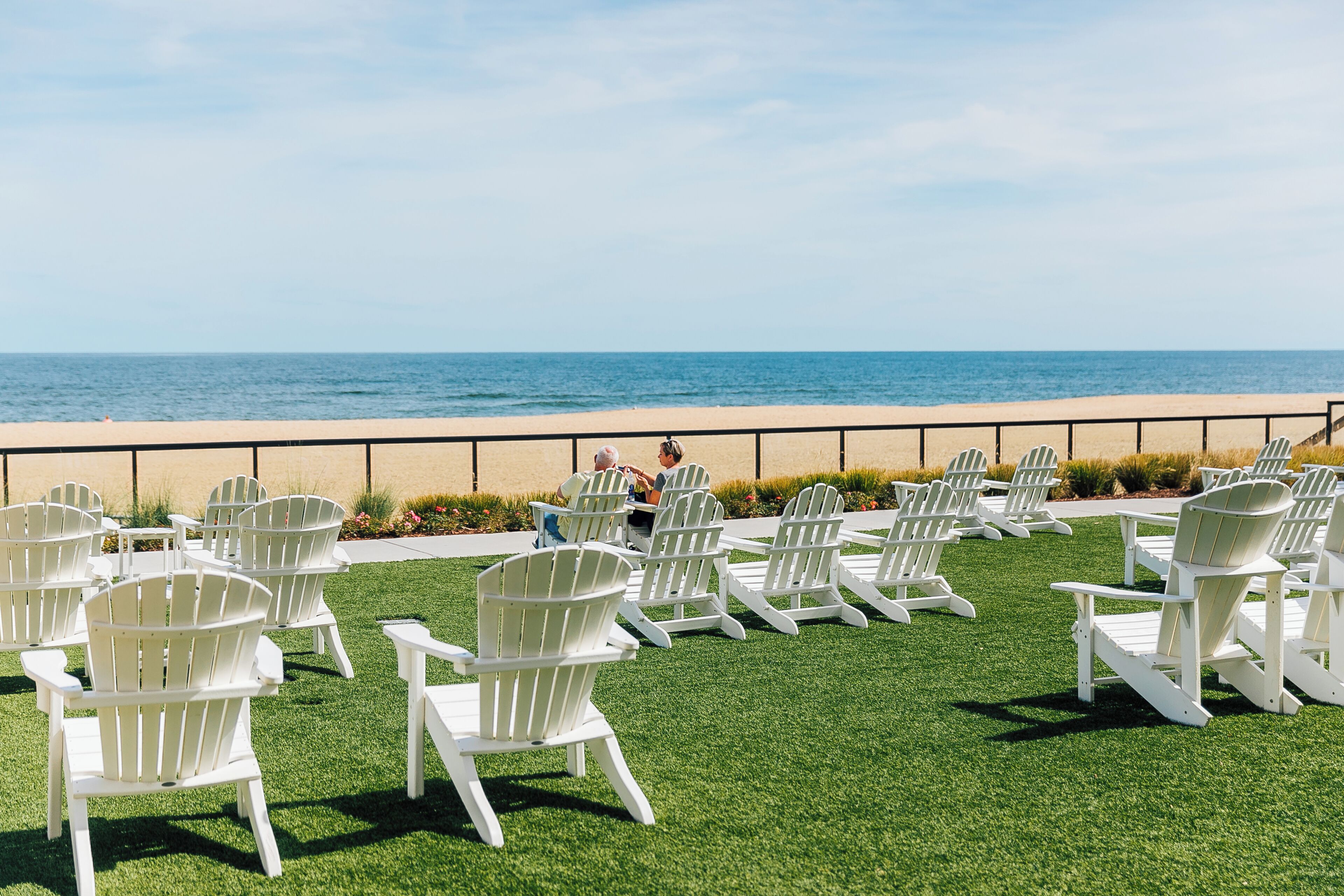 A beachside seating area with white Adirondack chairs arranged on artificial grass, overlooking the ocean.