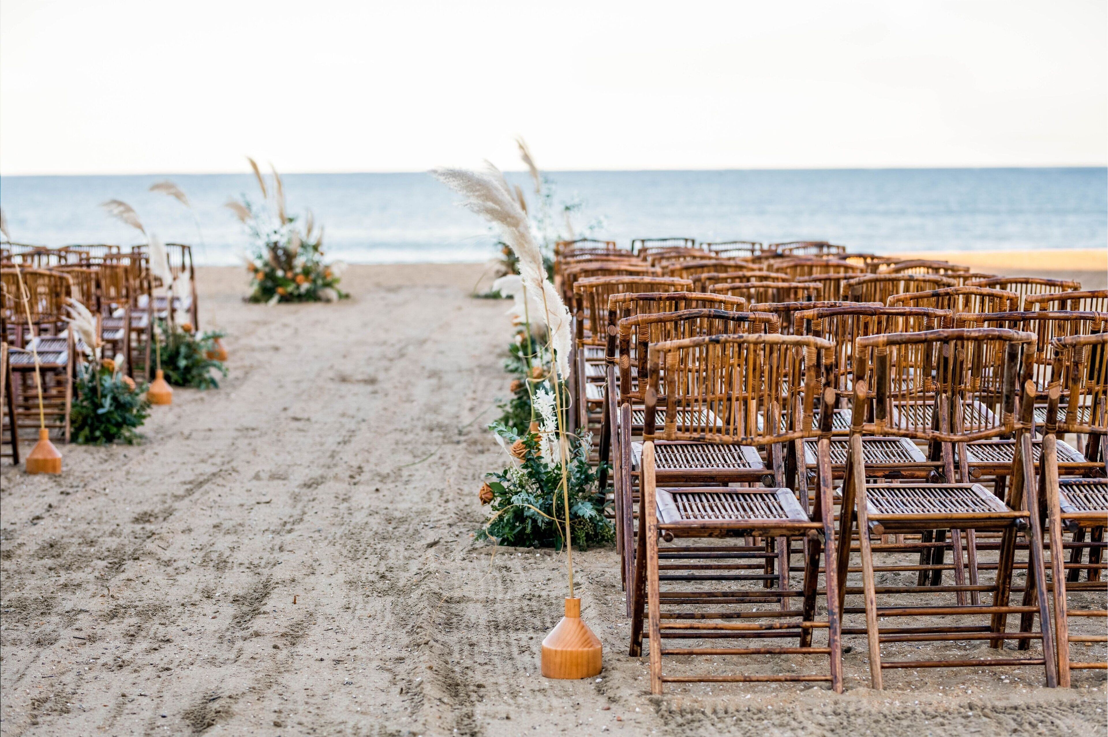 Rows of wicker chairs arranged on a sandy beach with a decorative floral arrangement and pampas grass.