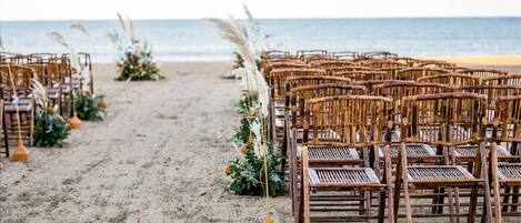 Filas de sillas de mimbre dispuestas en una playa arenosa, con un arreglo floral decorativo y pastos del pampa.