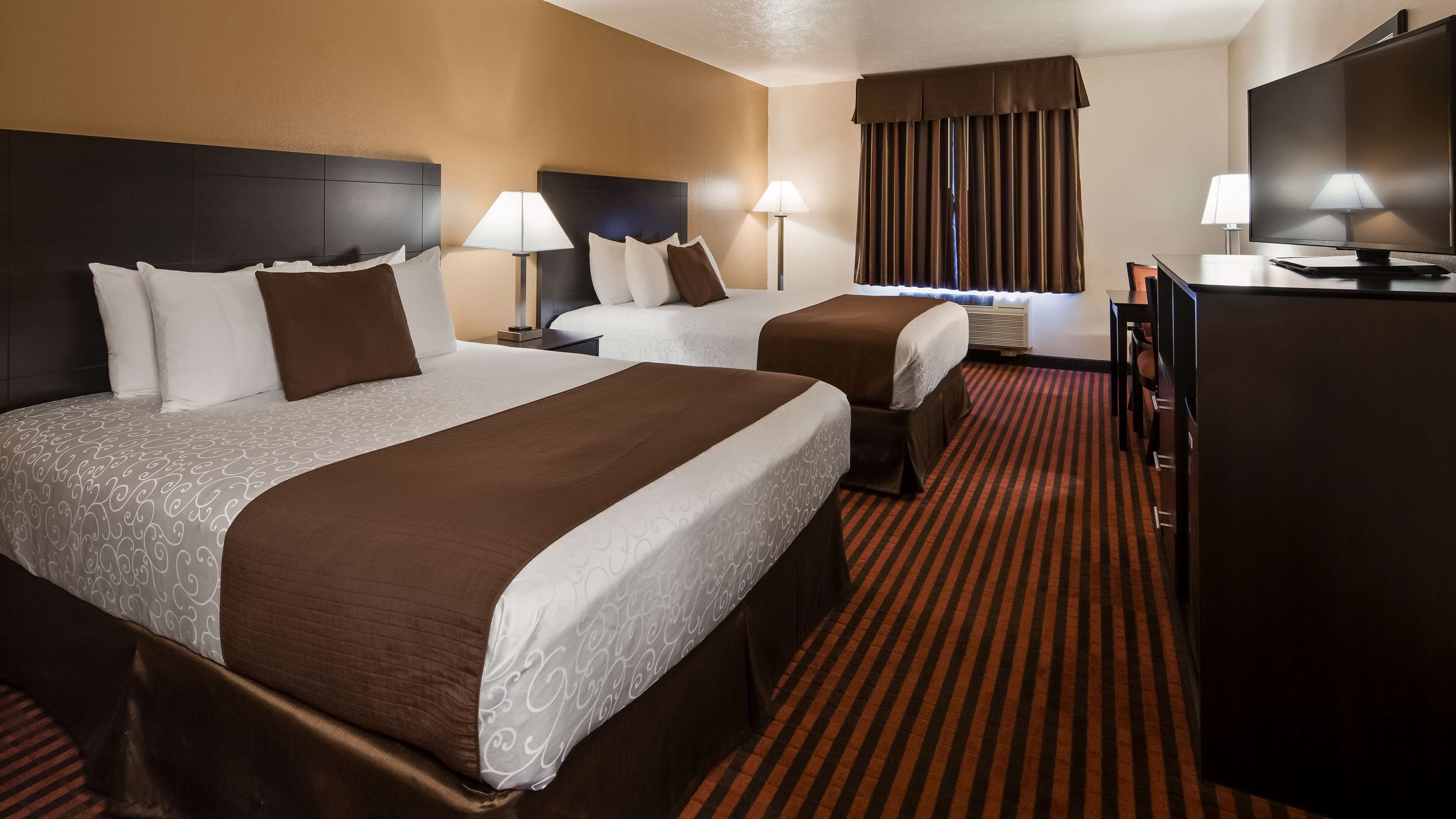 A hotel room with two beds, a desk, a television, and two lamps.